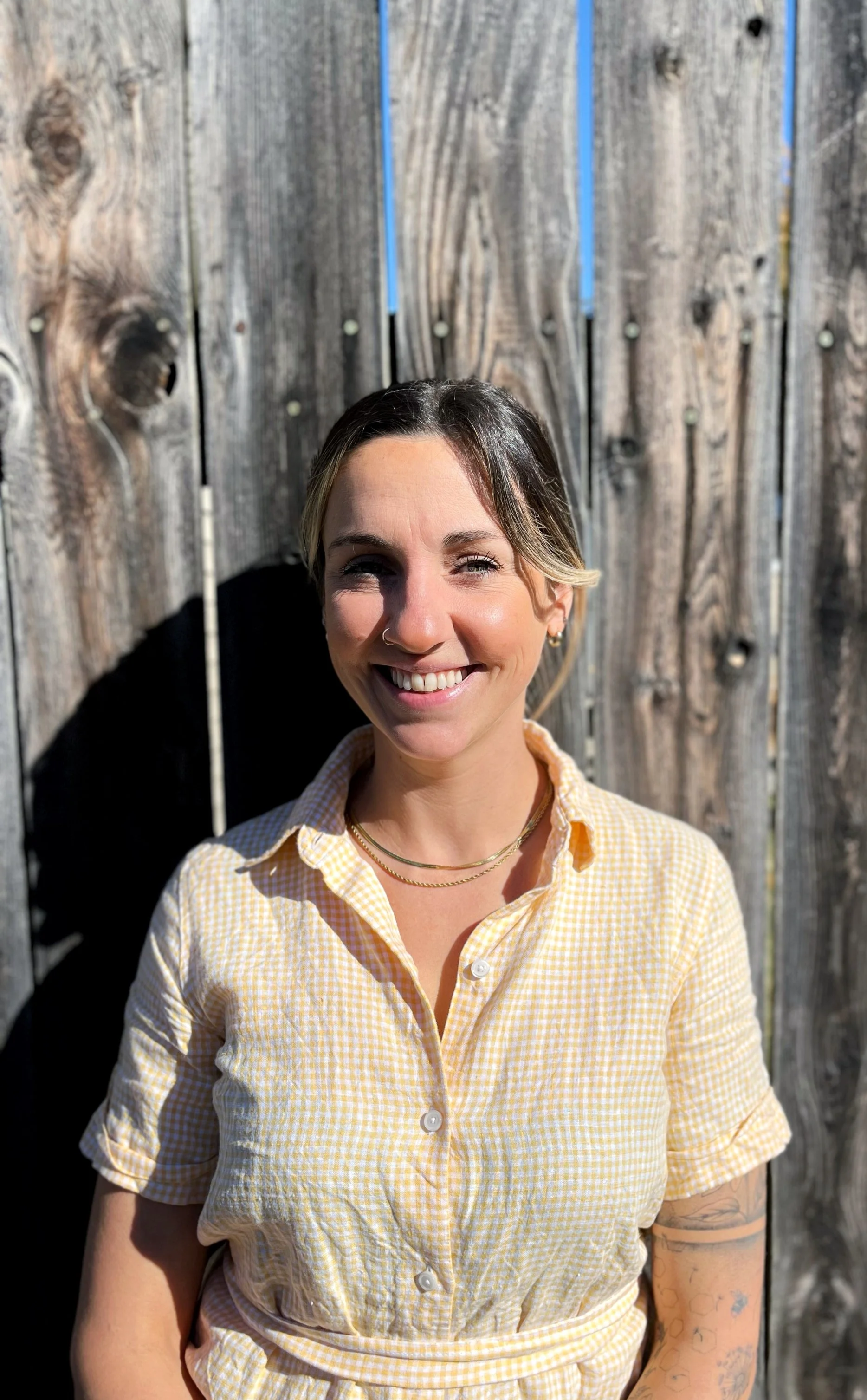 A woman named Riley is smiling outdoors in front of a weathered wooden fence, wearing a yellow checkered shirt and has tattoos on her full left arm. There is flowers and bees, mushroom and cedar all woven around it.
