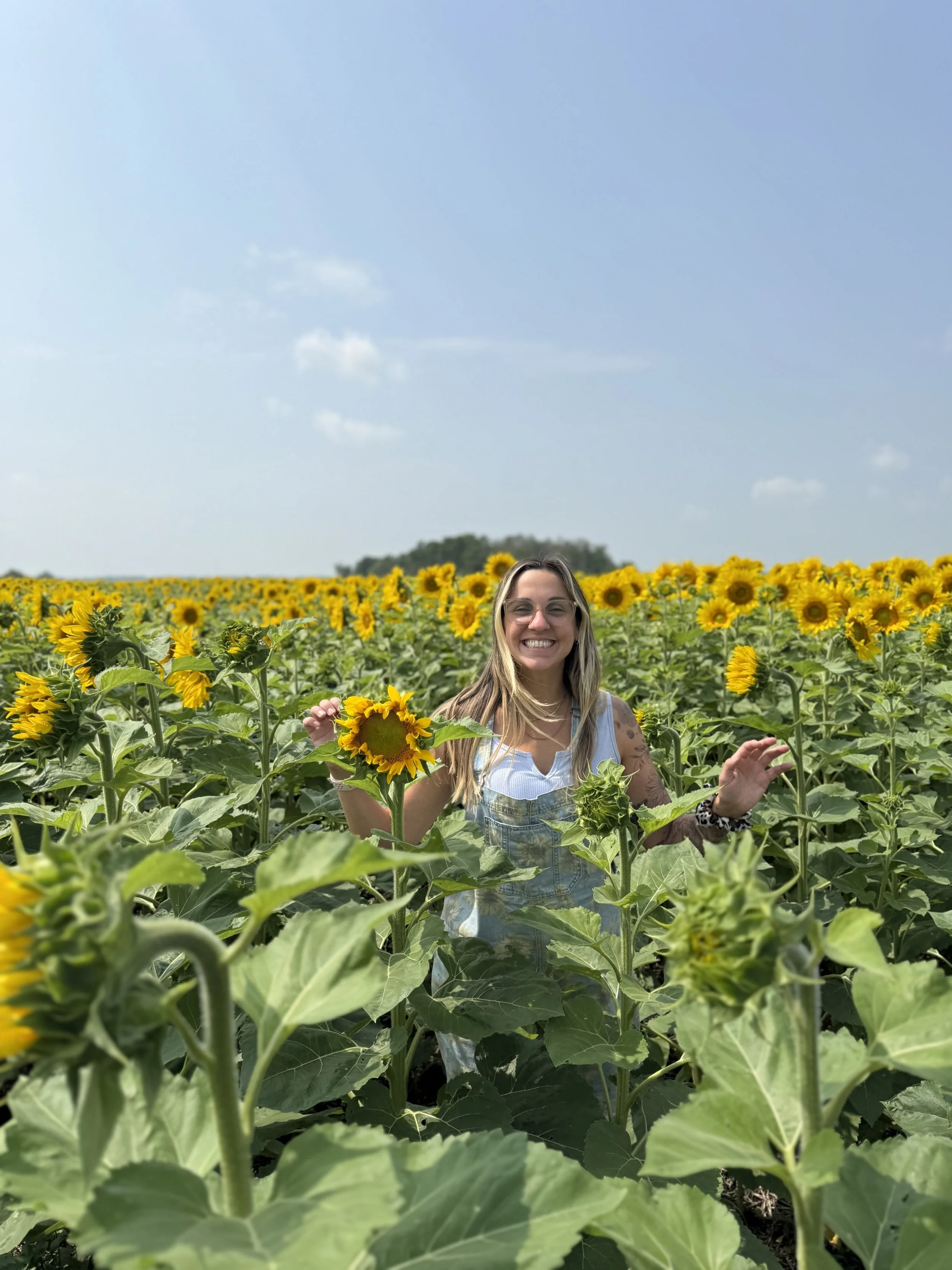 A woman named Riley smiling and holding a sunflower in a sunflower field under a blue sky with a few clouds.