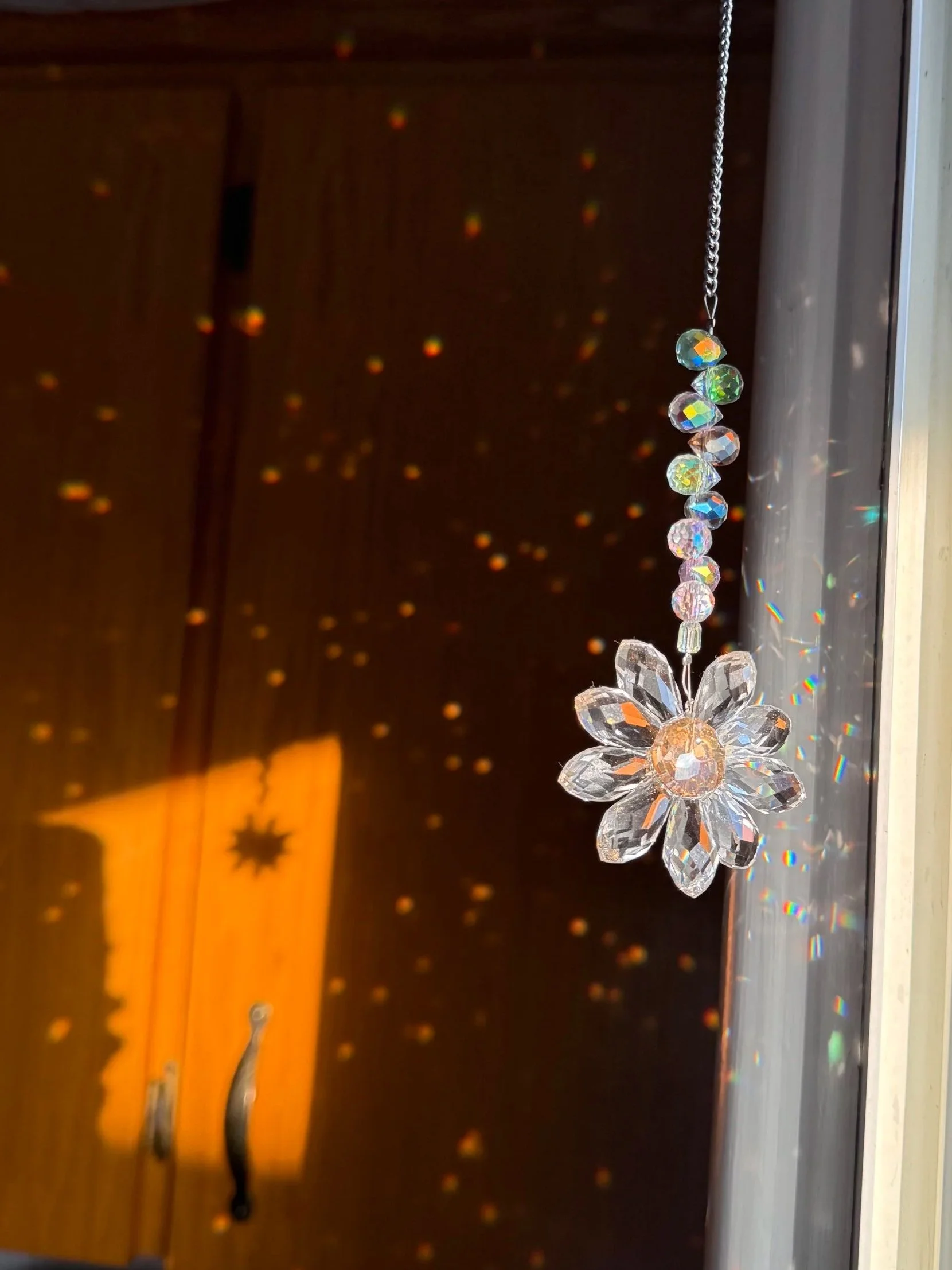 A hanging flower ornament with a silver chain against a wooden background decorated with small colorful light reflections from the sunlight hitting the crystal.