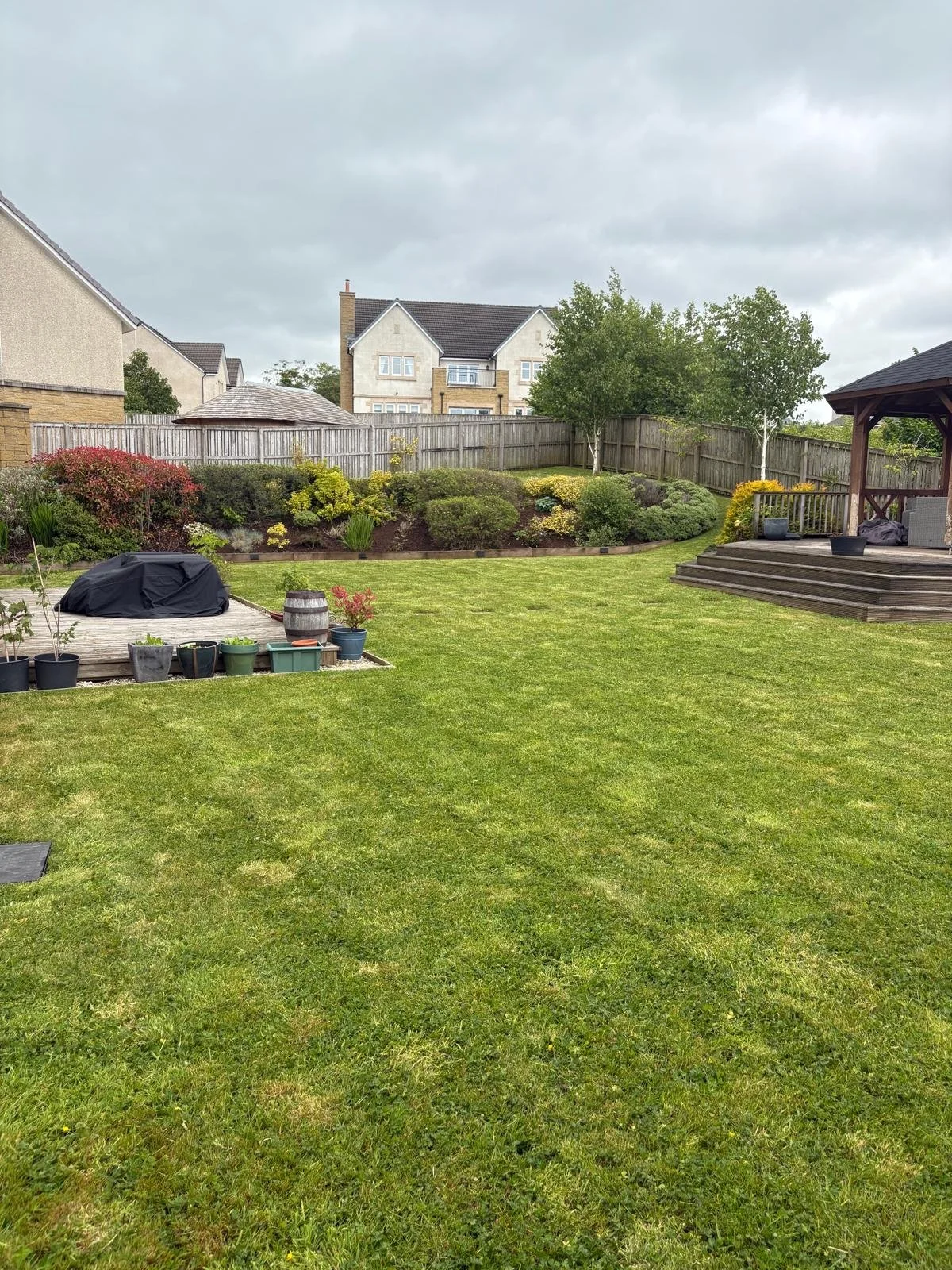 A backyard with a grassy lawn, a garden bed with bushes and flowers, a wooden deck with stairs, and potted plants. Houses and trees are in the background under a cloudy sky.