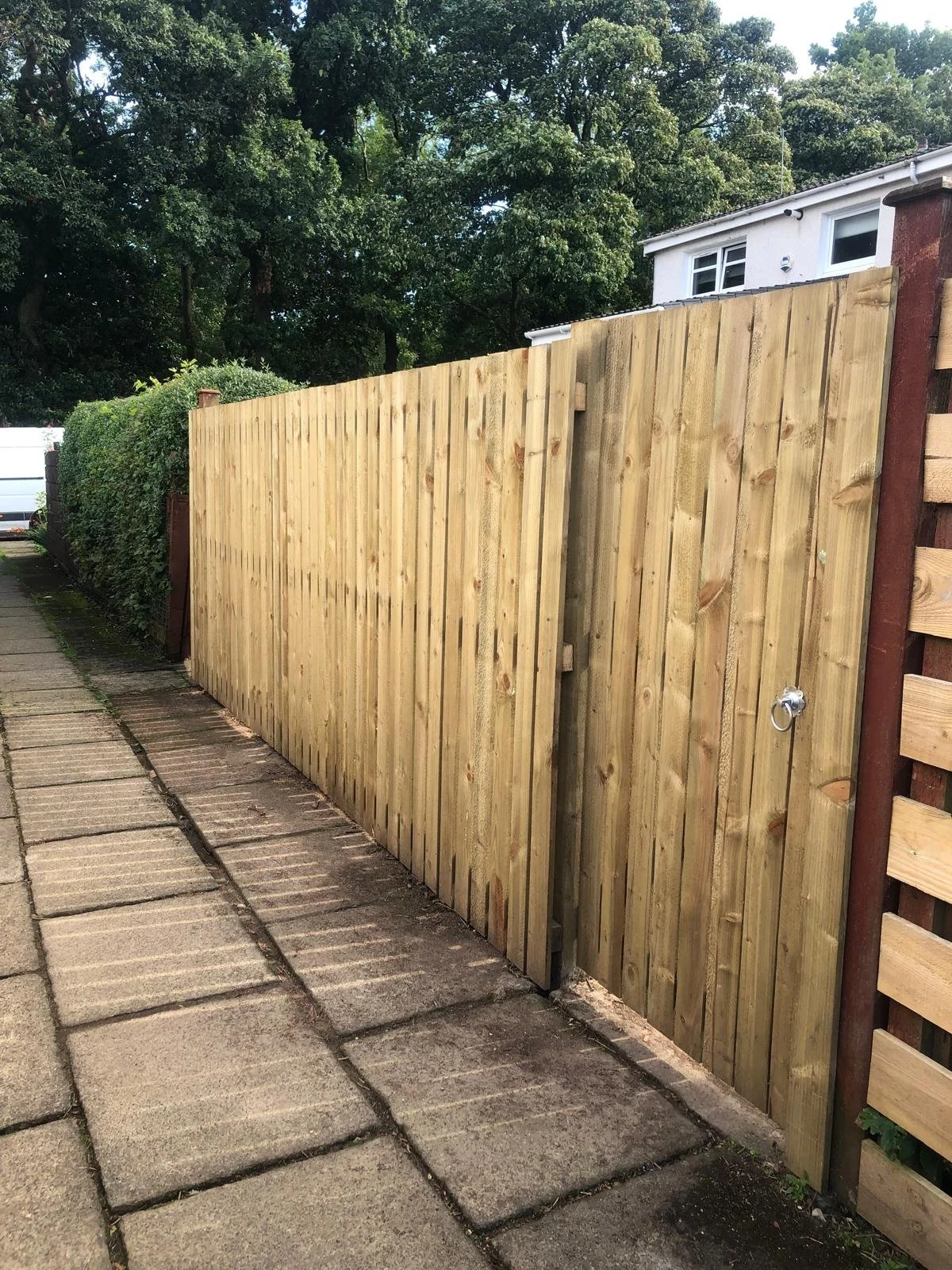 Newly built wooden gate and fence alongside a paved sidewalk with trees and a house in the background.