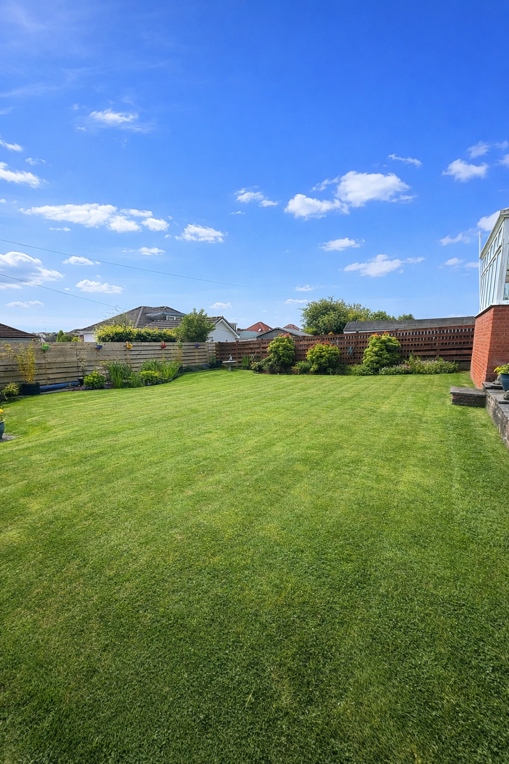 A lush green backyard with manicured lawn, a few small bushes, and flowering plants along a wooden fence under a bright blue sky with scattered white clouds.