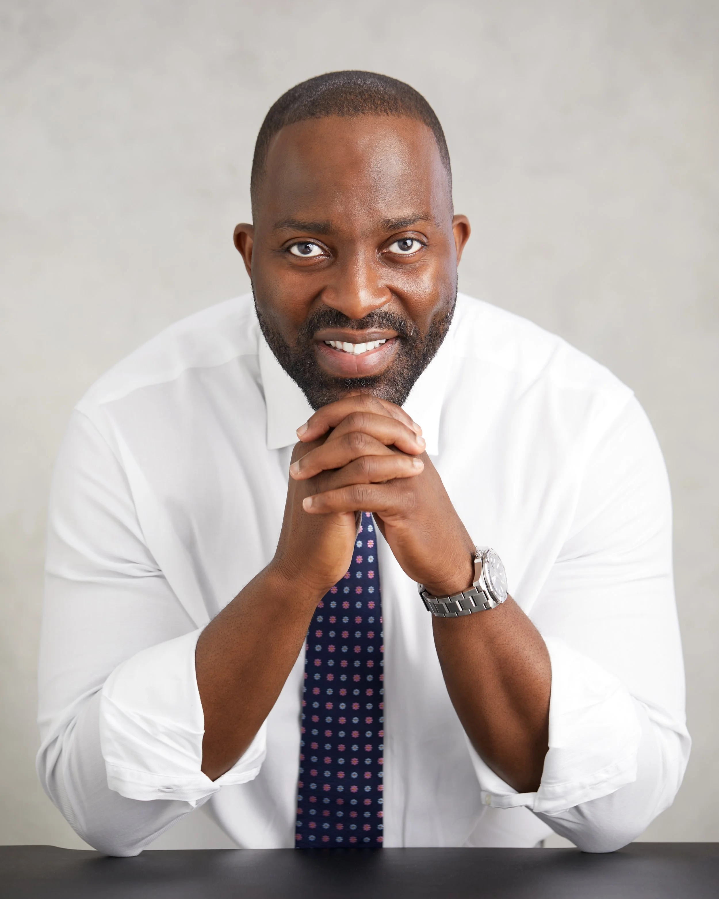 A man with a beard and short hair, wearing a white dress shirt, a patterned tie, and a watch, sitting with hands clasped.