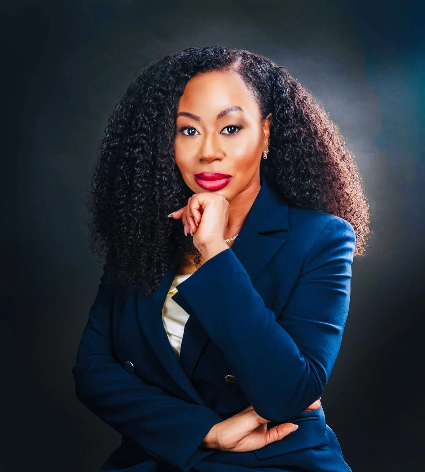 A woman with curly hair wearing a dark blue blazer and a light top, in a professional portrait pose against a dark background.