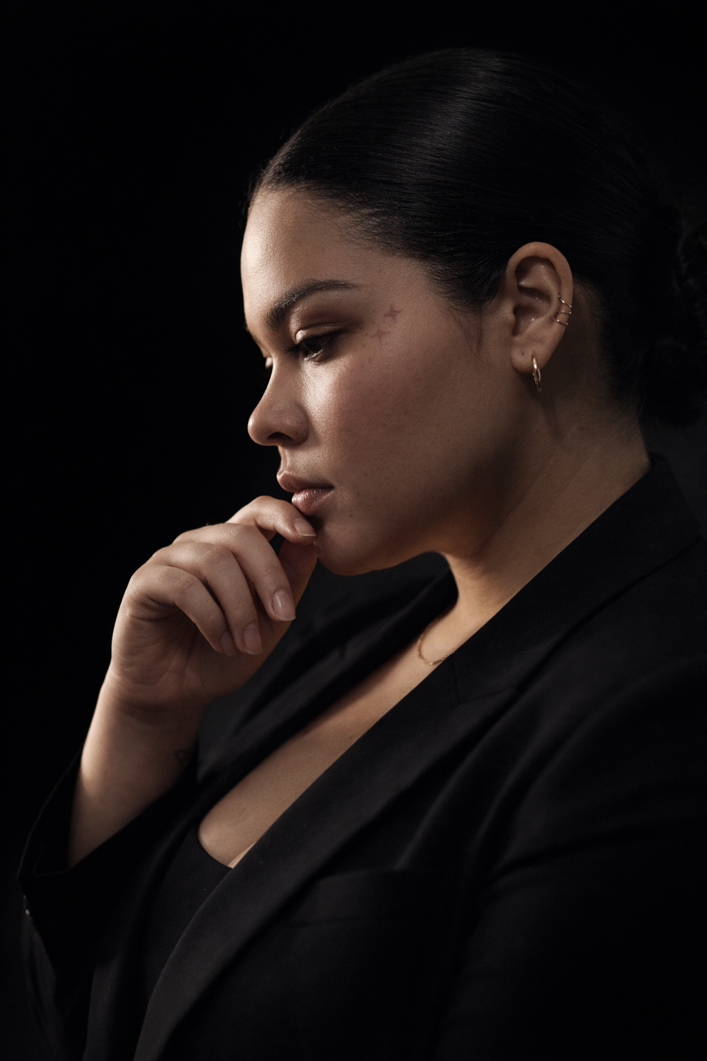 Side profile of a woman with dark hair, wearing black clothing, touching her chin thoughtfully, against a black background.