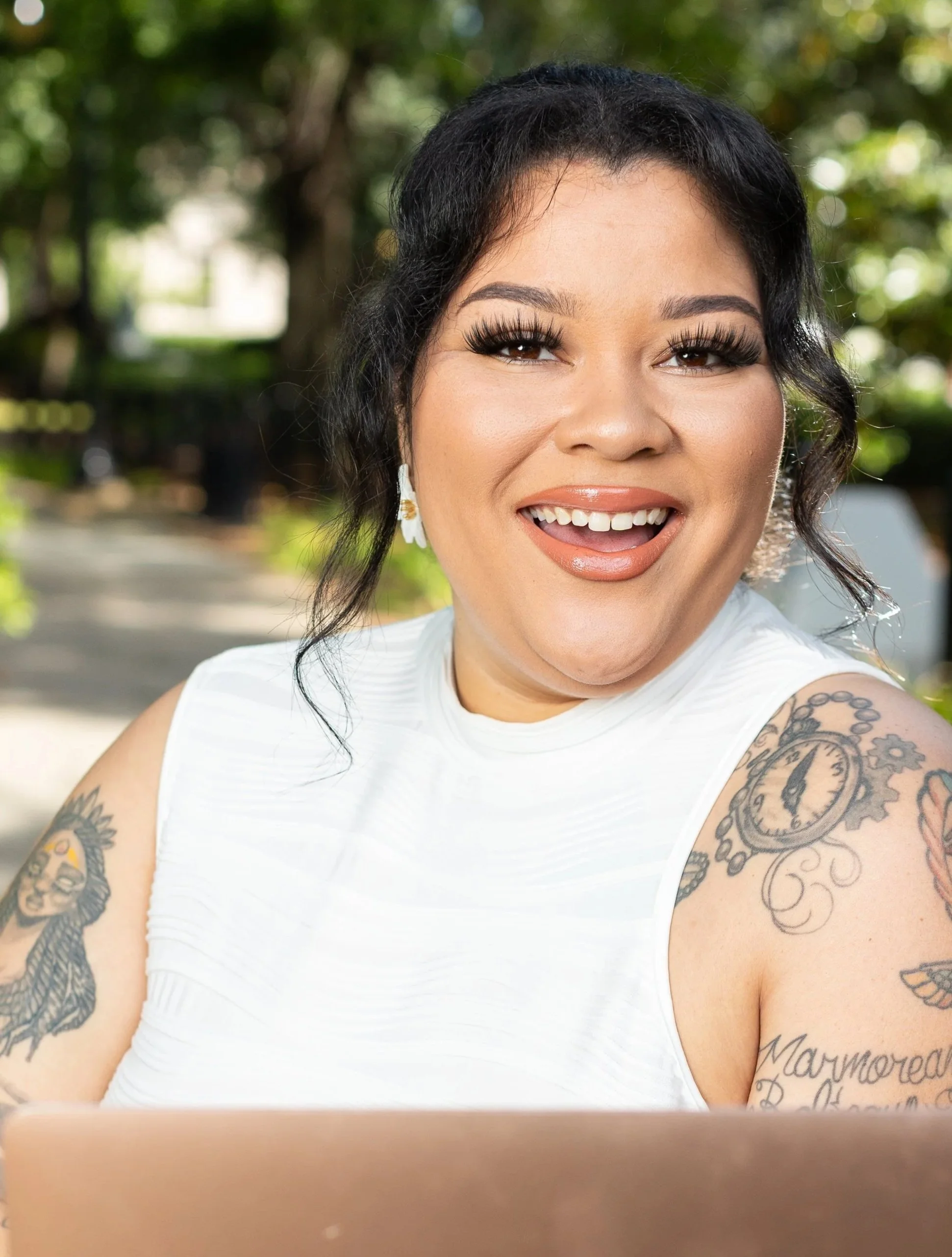 A woman with black hair, makeup, and tattoos on her arms is smiling outdoors, wearing a white sleeveless top, with greenery in the background.