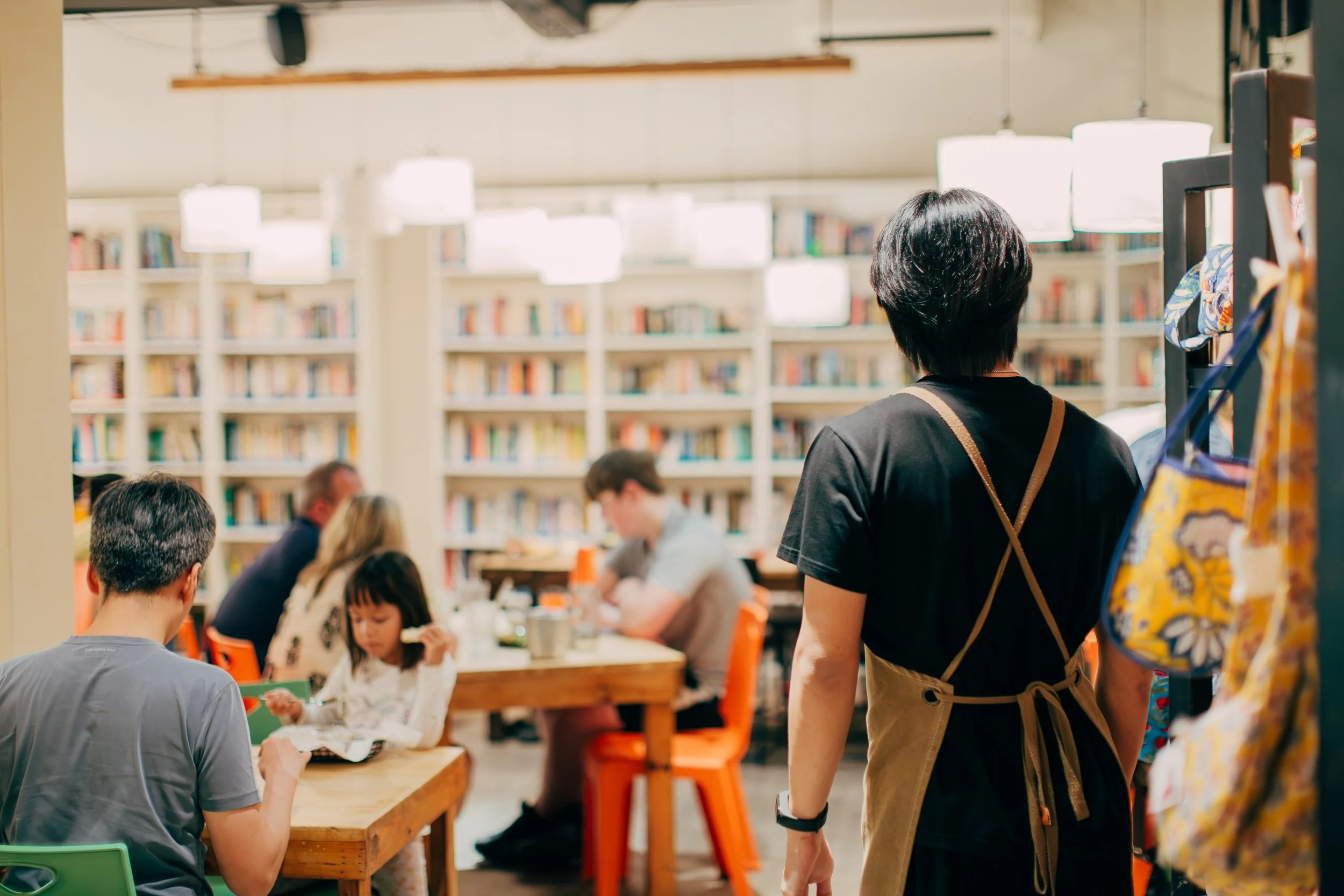 People gathered around tables in a bright library or bookstore with bookshelves in the background, one woman with short hair and a backpack observing the scene.