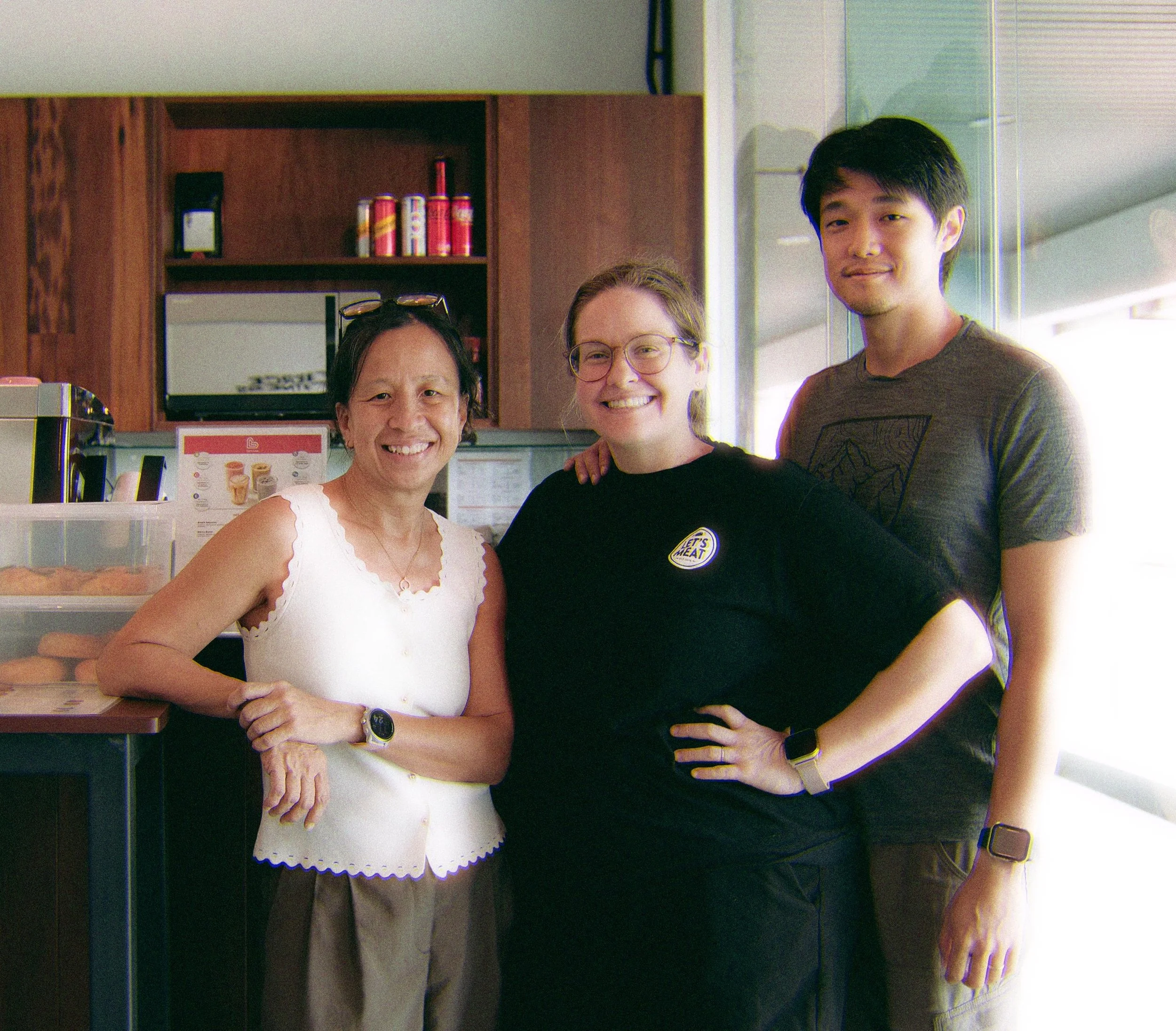 Four people standing together inside a store, smiling. From left to right: a middle-aged woman in a white sleeveless top, an older woman with glasses in a black top, a young woman with glasses, and a young man with black hair in a gray T-shirt.