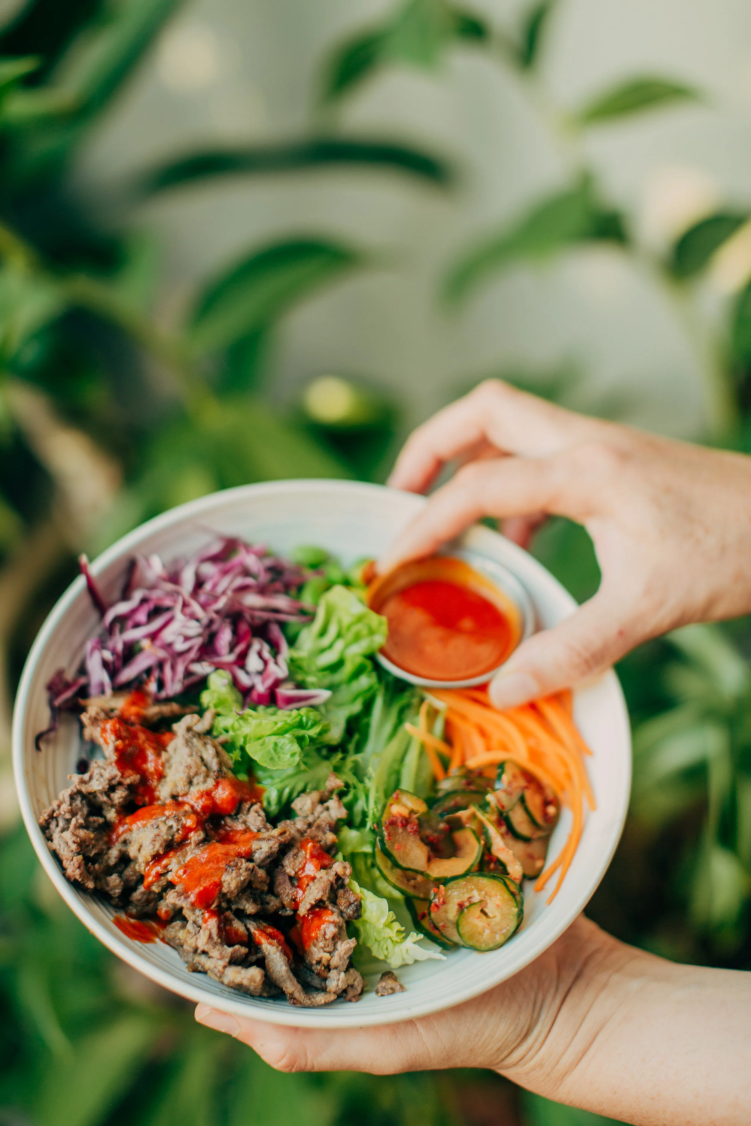 A hand holding a white bowl filled with various colorful vegetables and seasoned ground beef, with a blurred green leafy background.