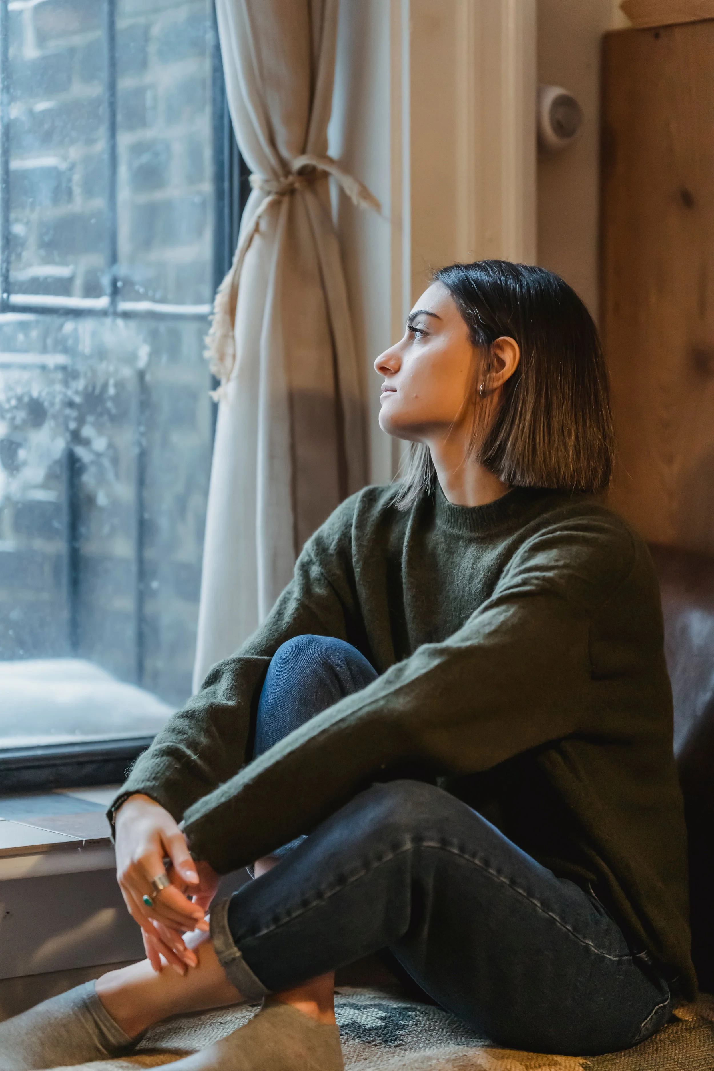 Woman sitting alone near a window looking thoughtful, representing emotional distance in family relationships