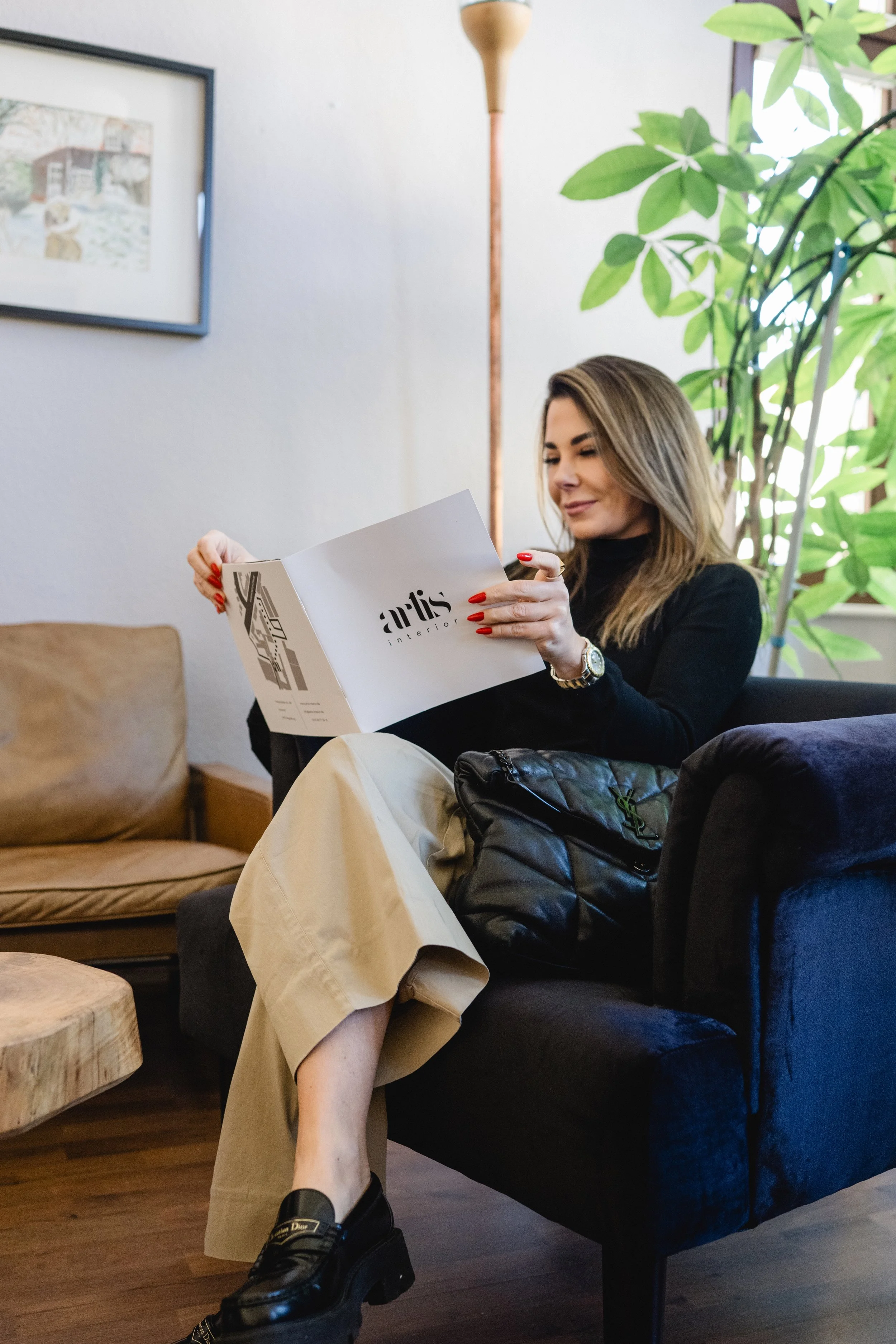 Eine Frau sitzt gemütlich auf einem schwarzen Sofa, liest eine Broschüre mit dem Titel 'arts interior' und trägt schwarze Schuhe, eine beige Hose und einen schwarzen Pullover. Im Hintergrund sind Pflanzen, ein Wandbild und ein Stehlampen, die den Raum warm wirken lassen.