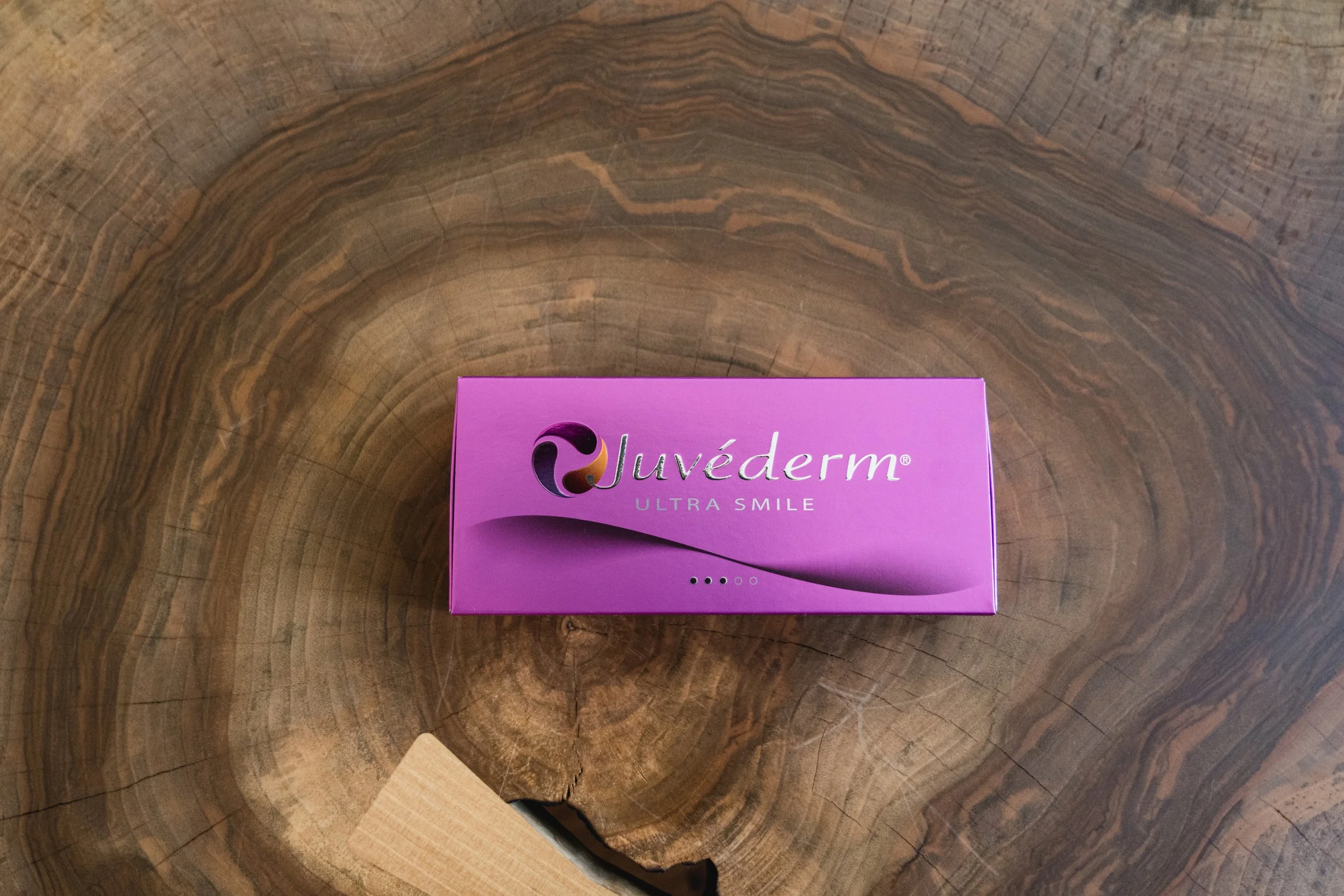 Eine pinke Verpackung mit dem Text 'Juvederm Ultra Smile' liegt auf einem braunen, gemaserten Holztisch.