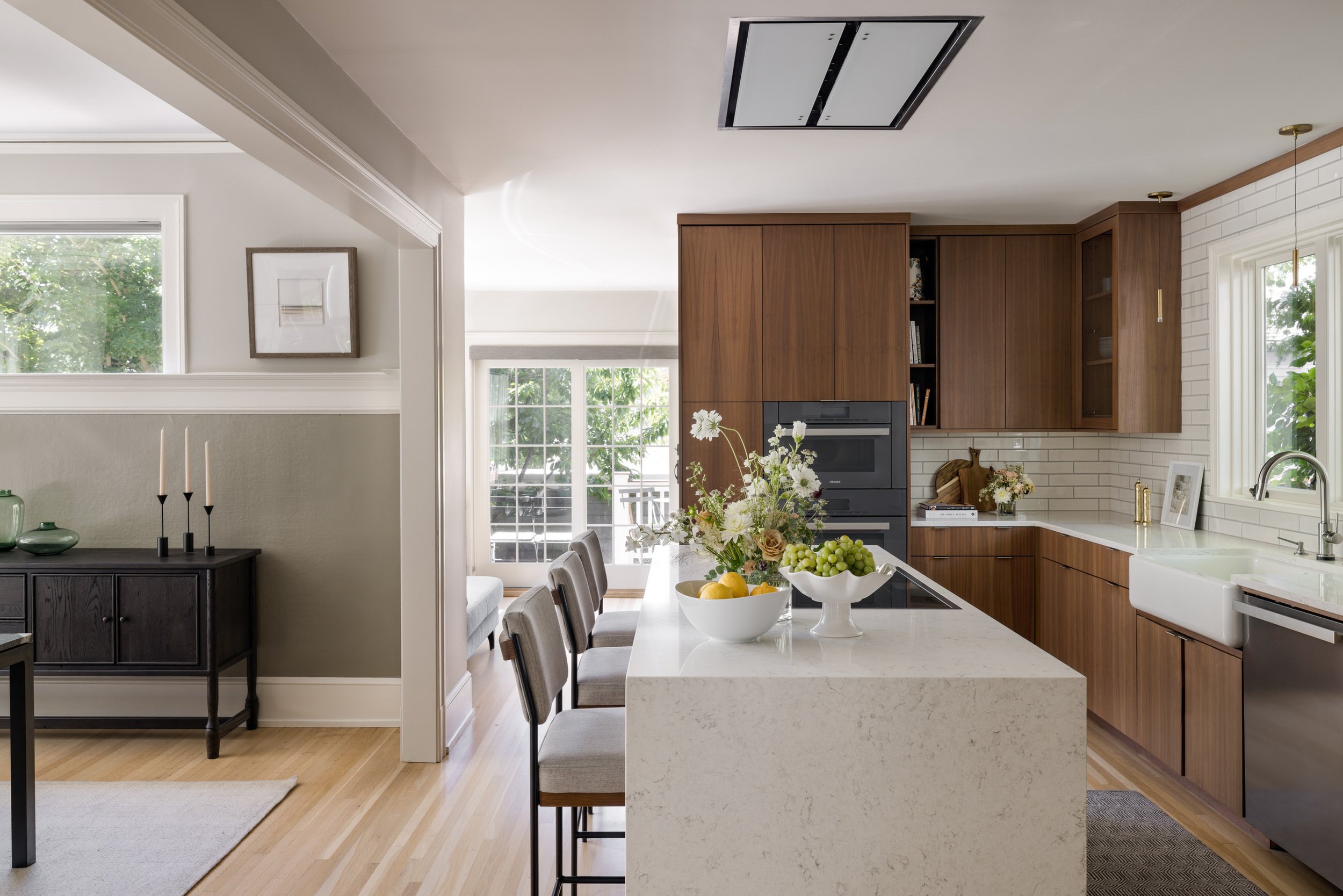 A bright kitchen with wooden cabinets, white subway tile backsplash, large windows, and a kitchen island with a flower arrangement and fruit bowls.