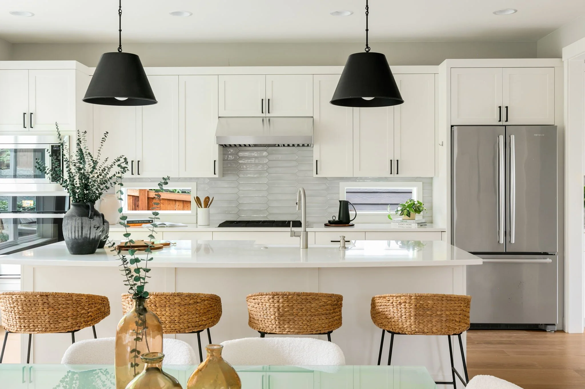 Modern white kitchen with hanging black pendant lights, stainless steel refrigerator, and a kitchen island with woven rattan chairs and decorative vases.