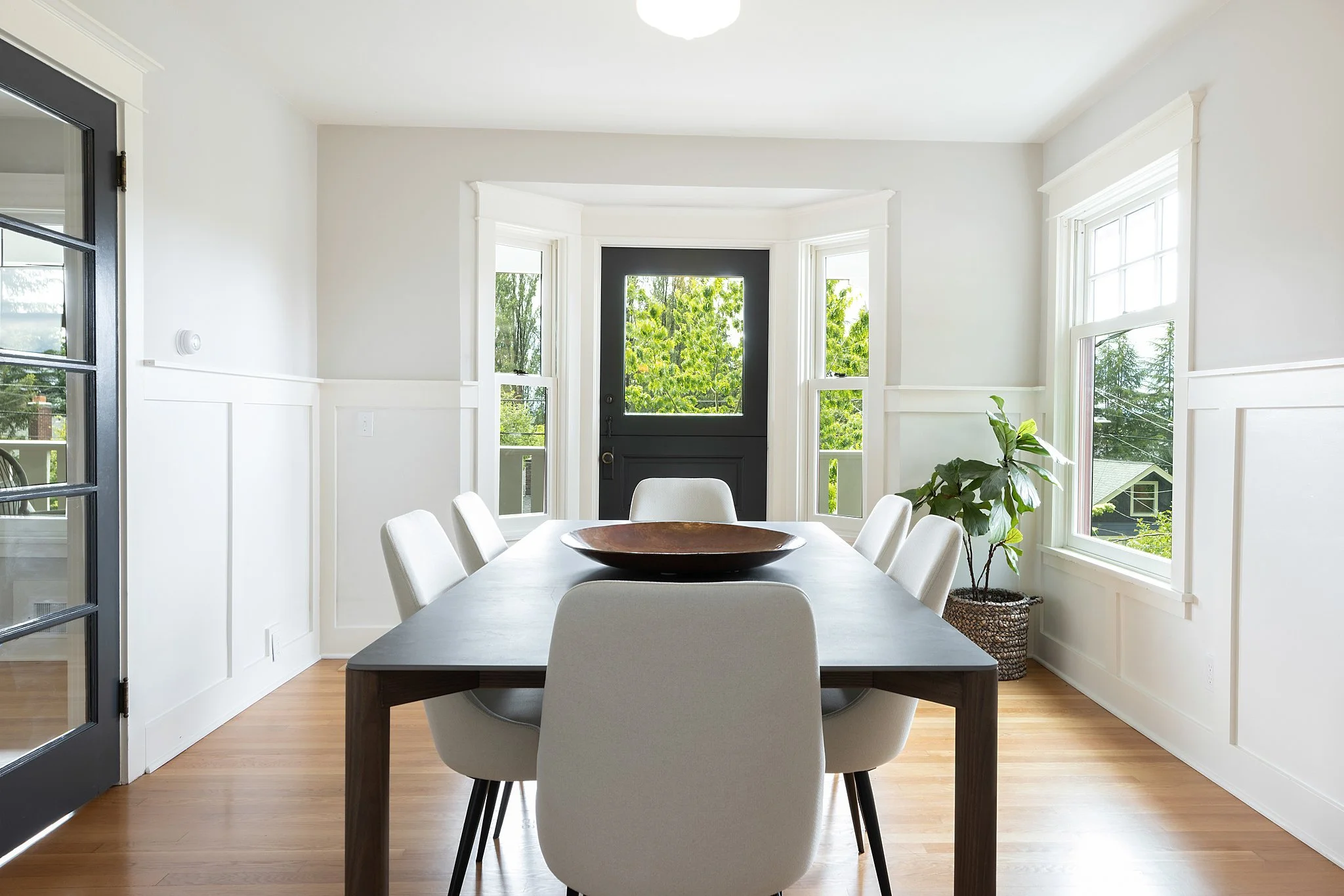 Dining room with a black front door, white walls, large windows with views of green trees, a wooden dining table, white upholstered chairs, and a potted plant.