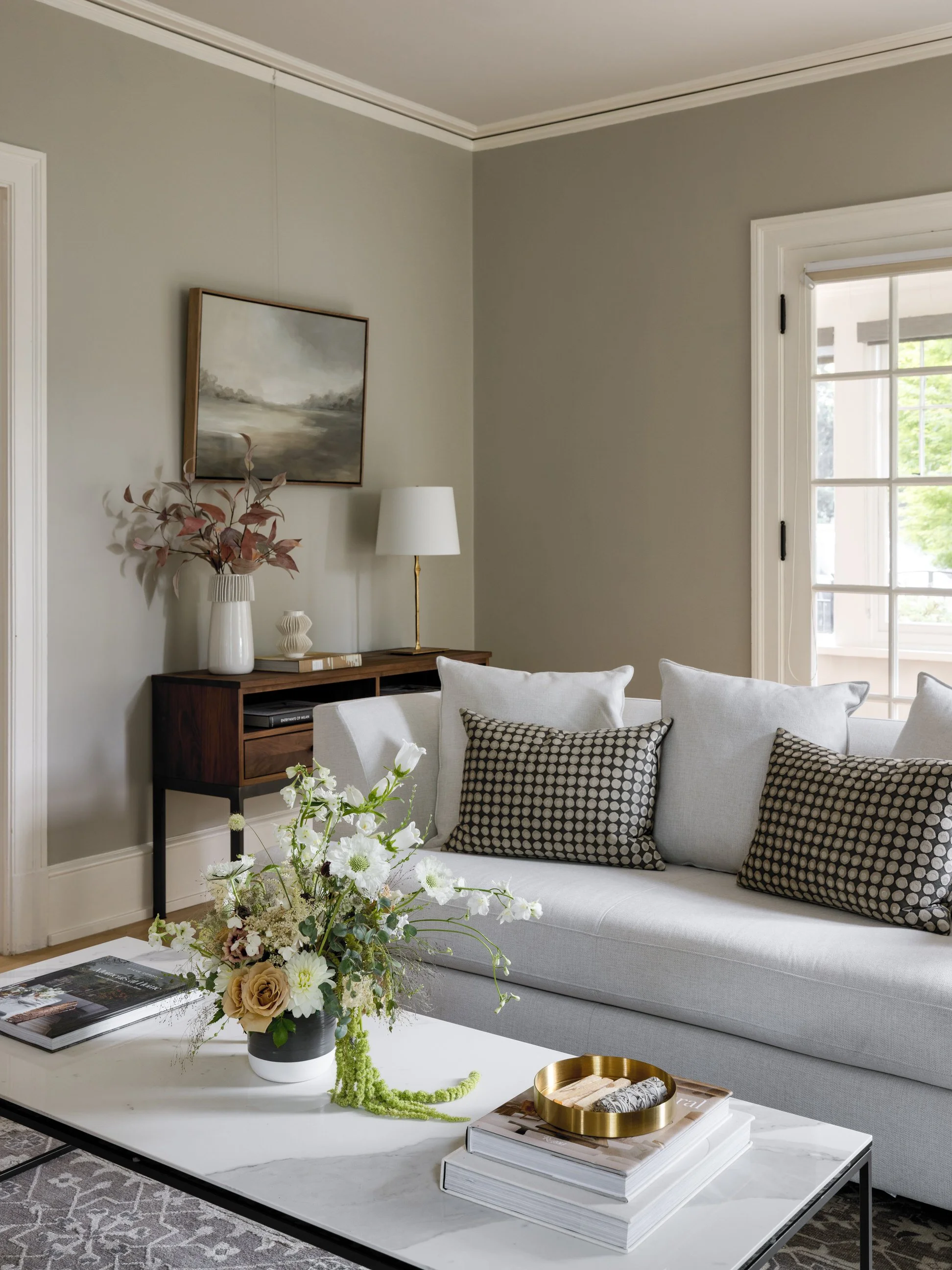 A living room with a white sofa adorned with black and white patterned throw pillows, a marble coffee table with a large floral arrangement, a wooden sideboard with a white vase and a table lamp, a wall painting, and a window with white trim letting 