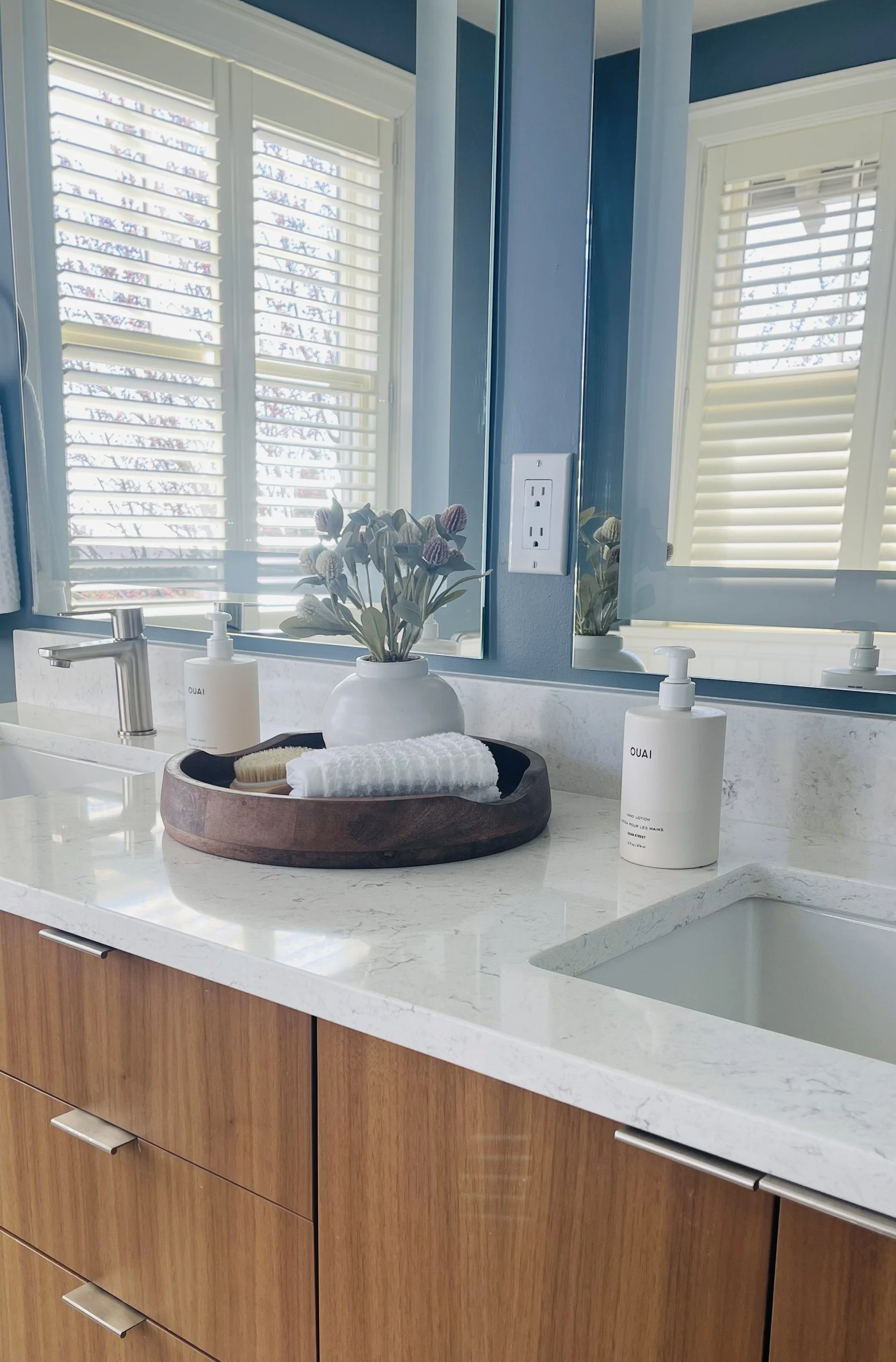 Bathroom countertop with a white soap dispenser, a white lotion bottle labeled 'OUAI', a white towel, a small brush, a potted plant, and a large mirror reflecting windows with white shutters and a blue wall.