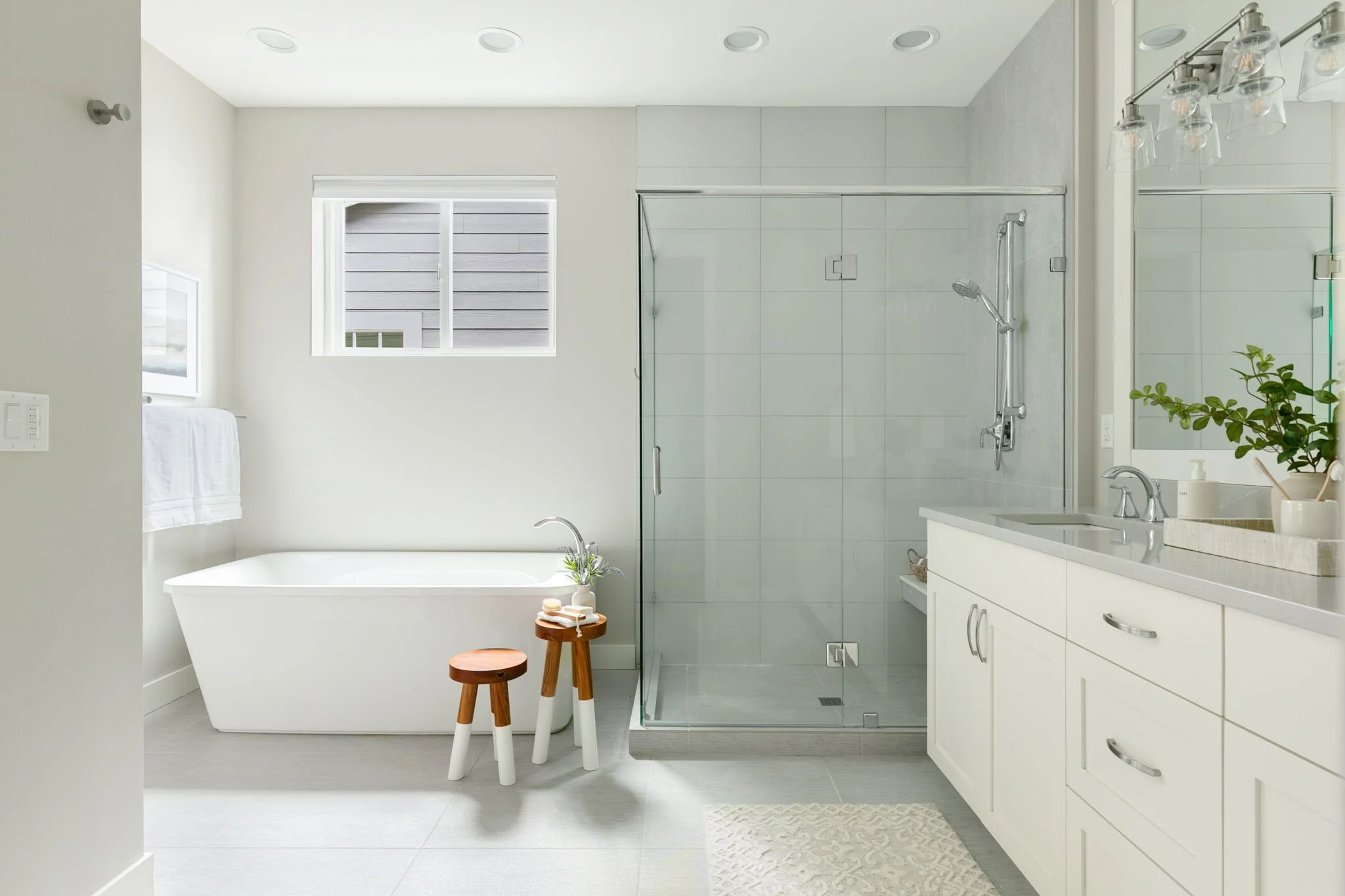 Modern white bathroom with a bathtub, a glass-enclosed shower, white vanity with a sink, and small stools, decorated with green plants and having two windows.