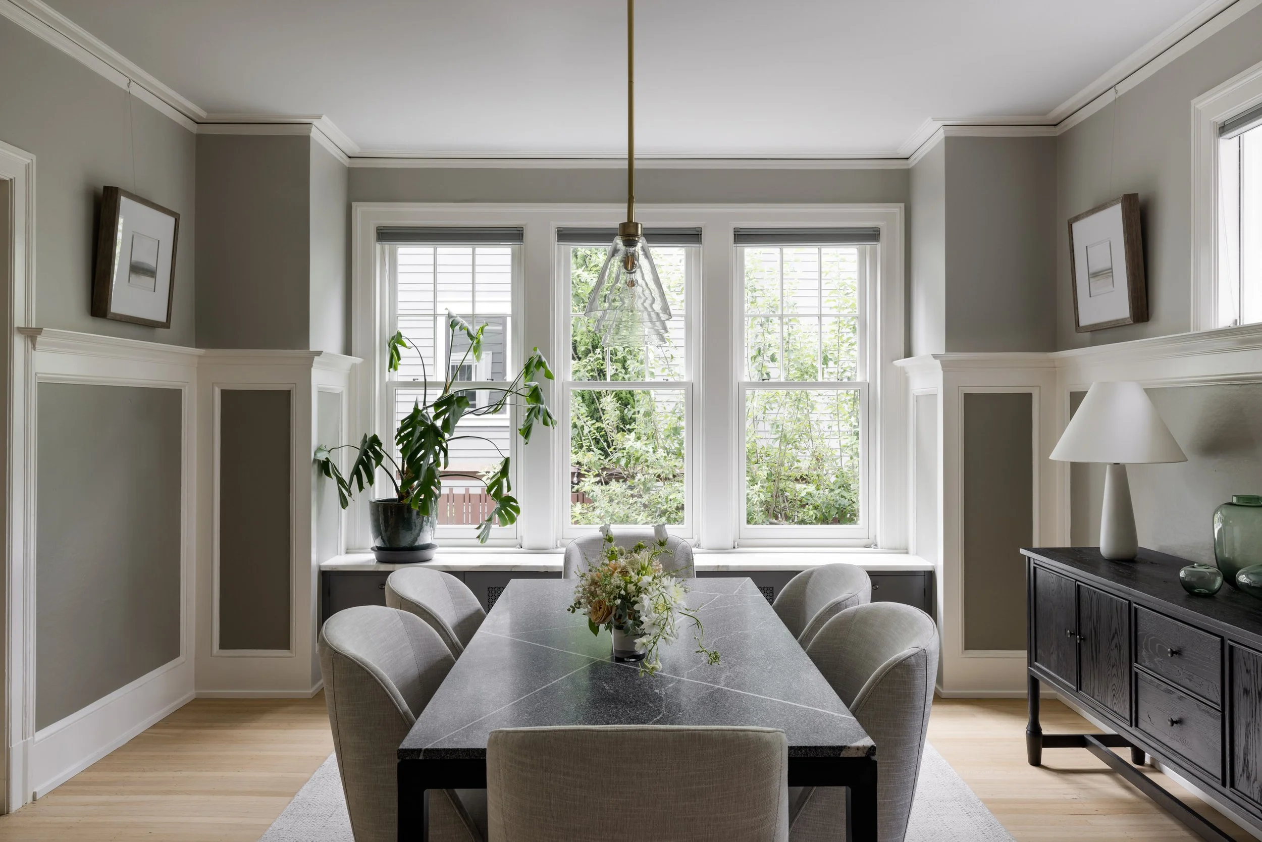 A dining room with large windows, a vase with flowers on the table, a plant near the window, wall art, and a cabinet with decorative items.