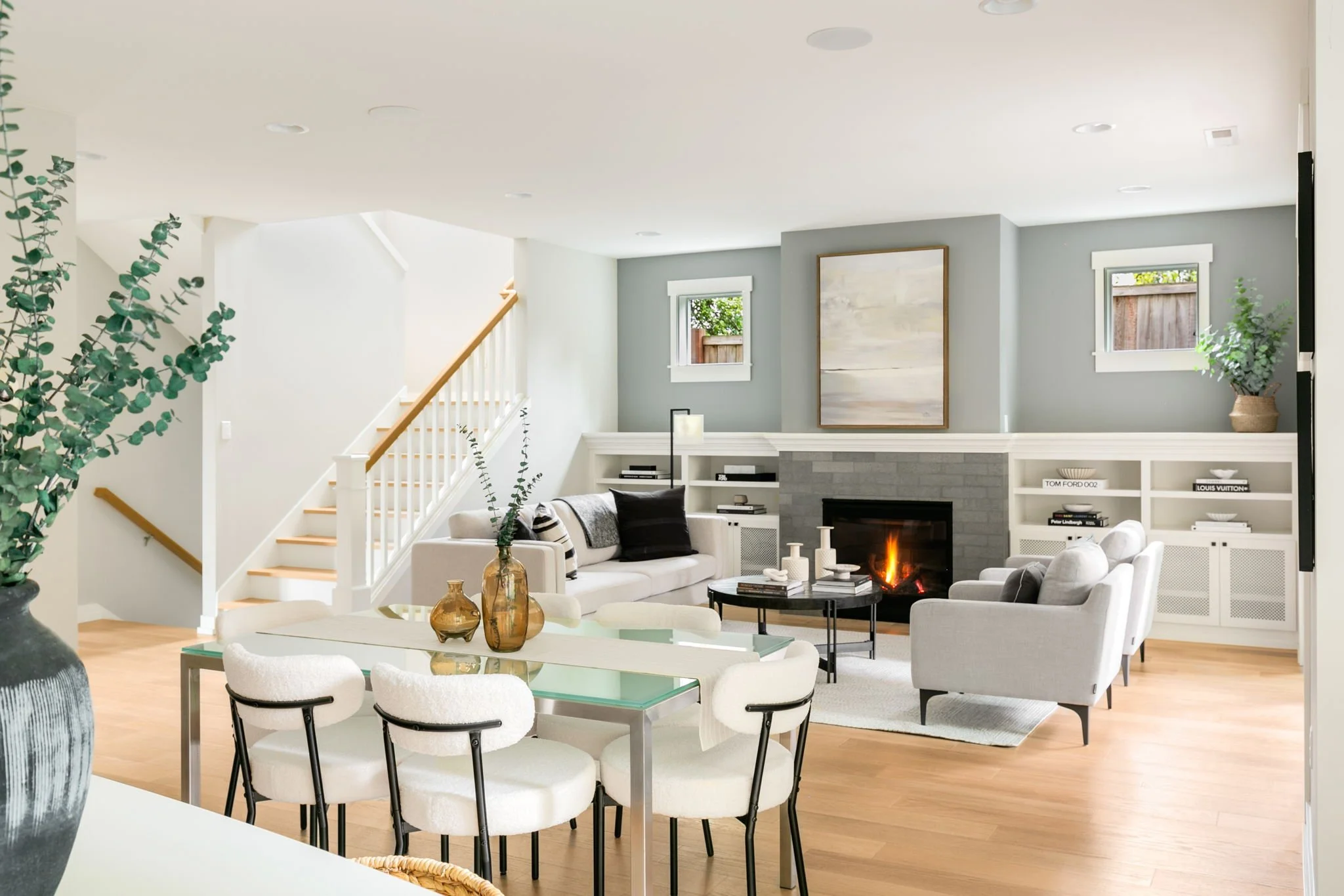 Open-concept living and dining room with modern furniture, a fireplace, and decorative elements, featuring large windows and light-colored walls.