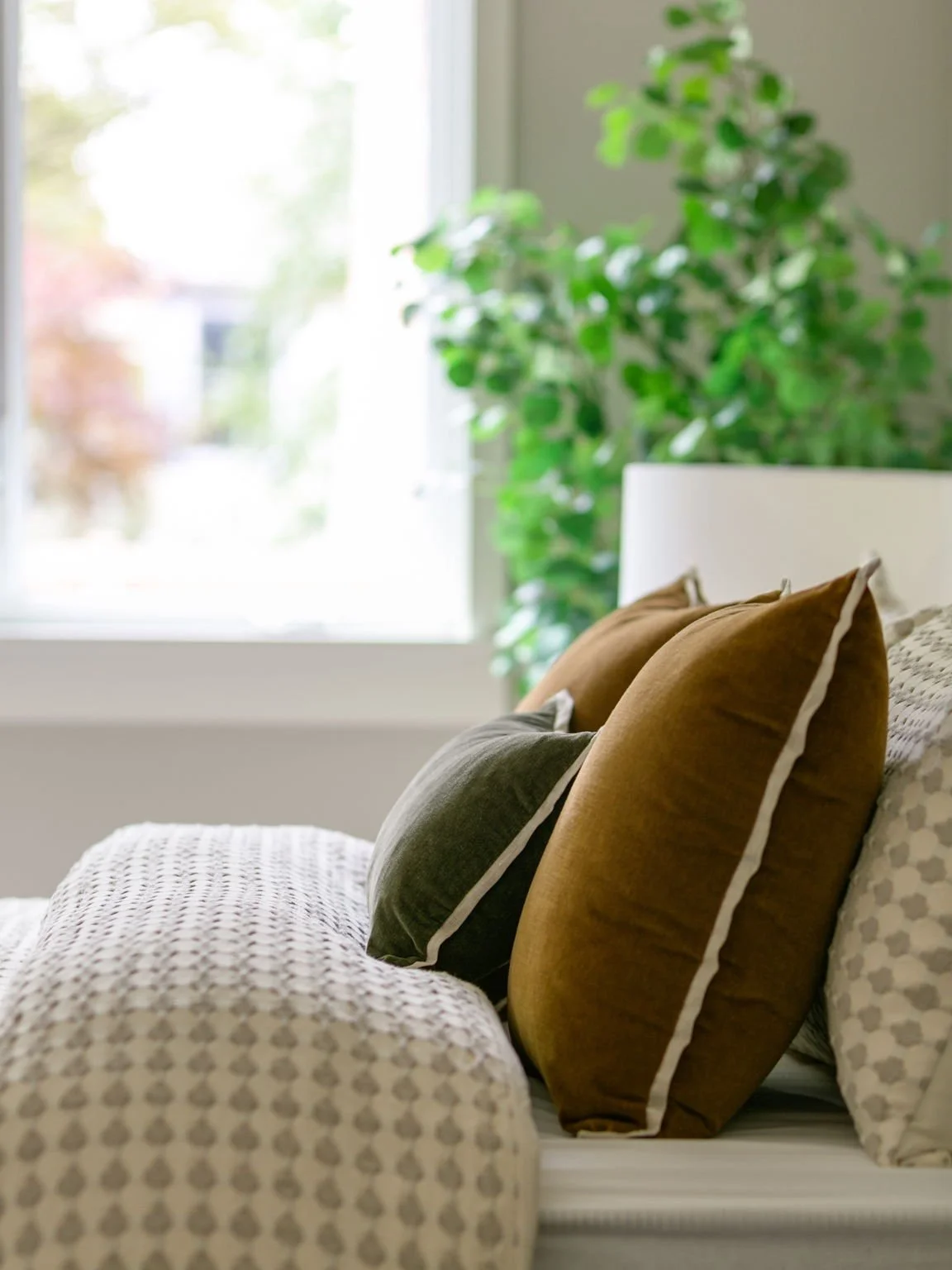 A cozy bedroom with a white bed frame, beige and brown pillows, a patterned blanket, and a green plant near a window with natural light.