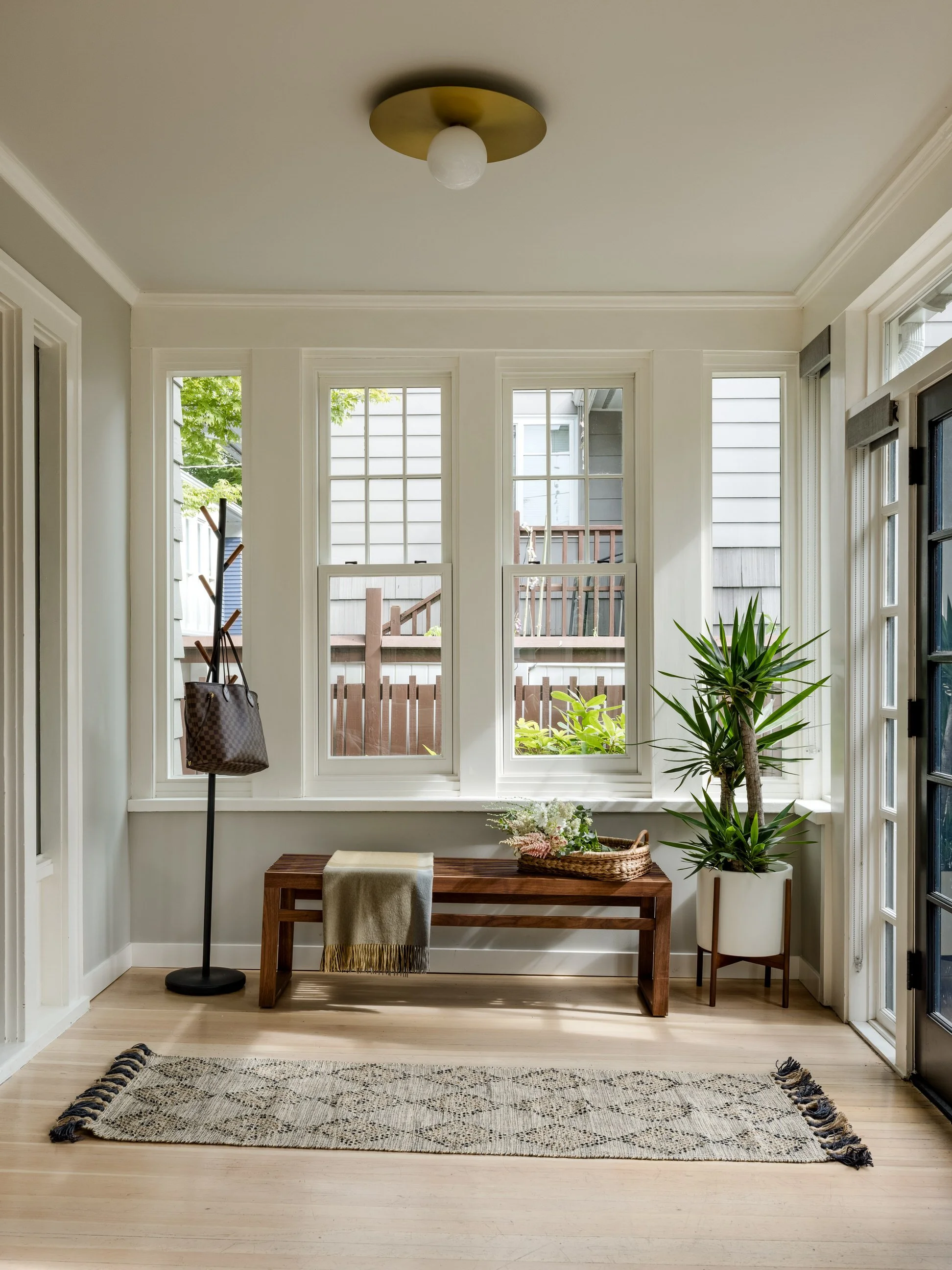 A bright room with a large window showcasing a backyard with trees and a wooden fence. The room features a wooden bench with a blanket, potted plants, and a standing coat rack.