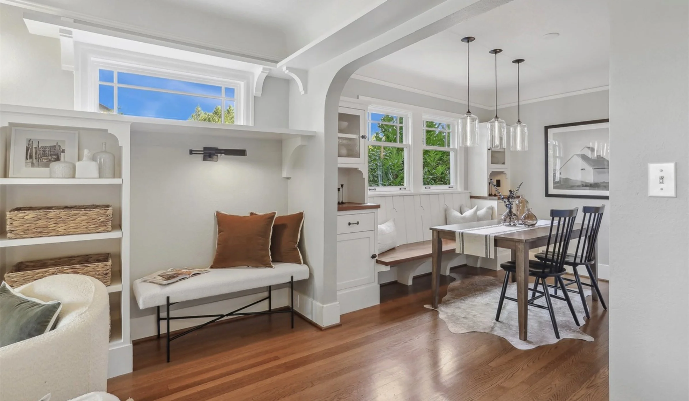 Cozy living room with white walls, large windows showing greenery outside, hanging light fixtures, and a wooden dining table with black chairs.