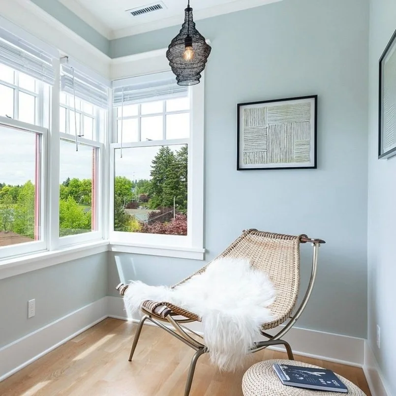 A corner of a bright room with large windows showing a view of greenery outside. There is a modern woven chair with a furry white throw and a stool with a magazine. Decorative framed art hangs on white walls, and a black woven pendant light hangs fro