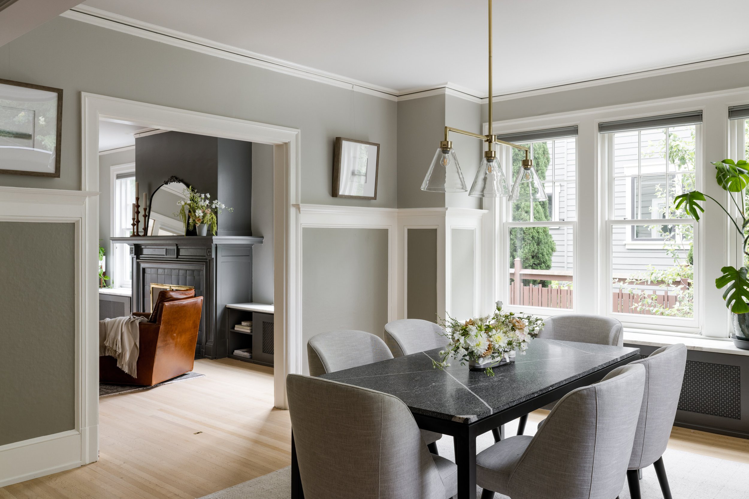 Living and dining room area with large windows, light gray walls, white trim, a black dining table with gray chairs, a chandelier, and decorative plants.