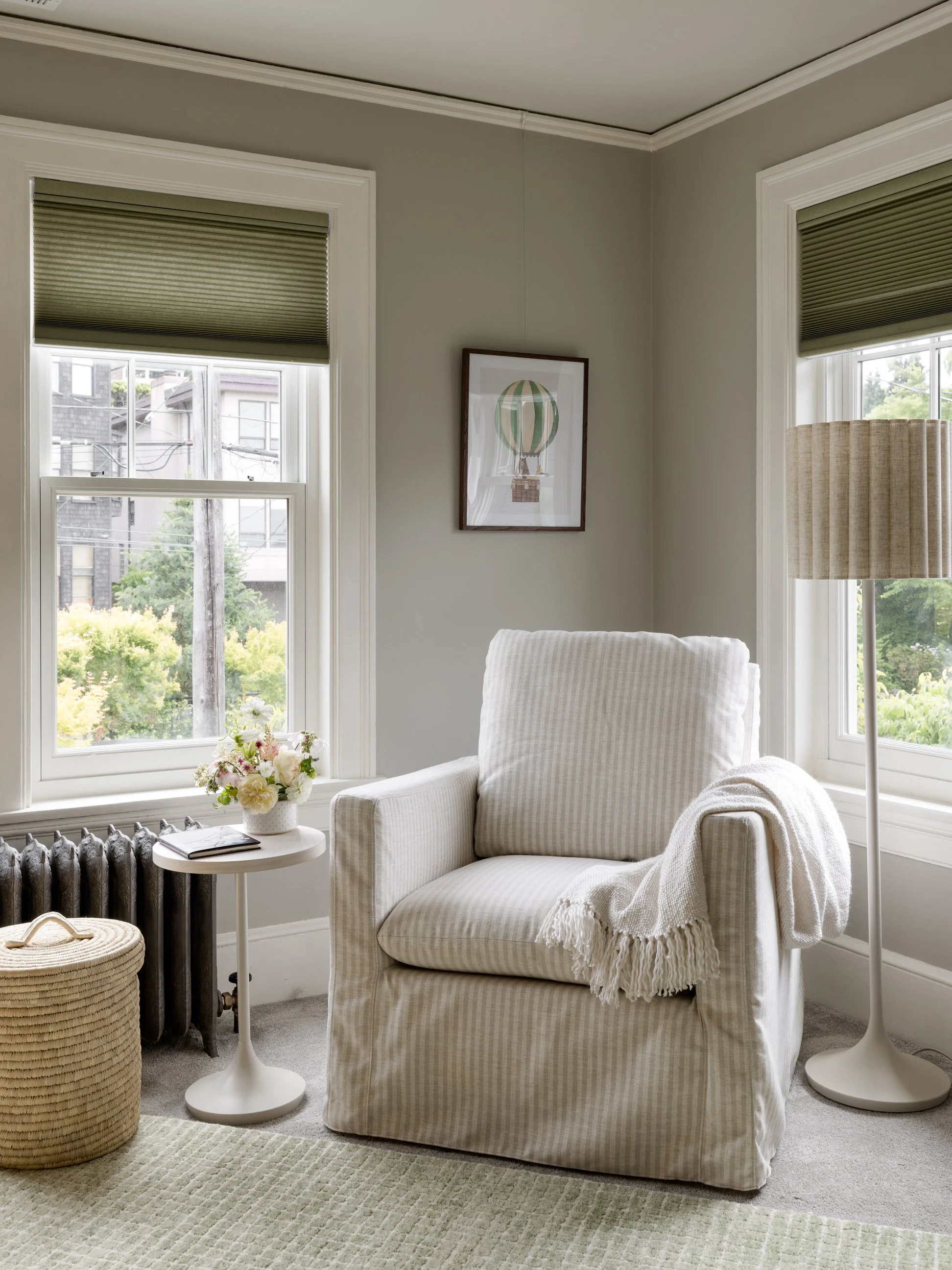 A cozy living room corner featuring a white armchair with a striped fabric pattern, a cream throw blanket, and a small side table with a floral arrangement and a book. Two large windows with green blinds allow natural light, and a framed hot air ball