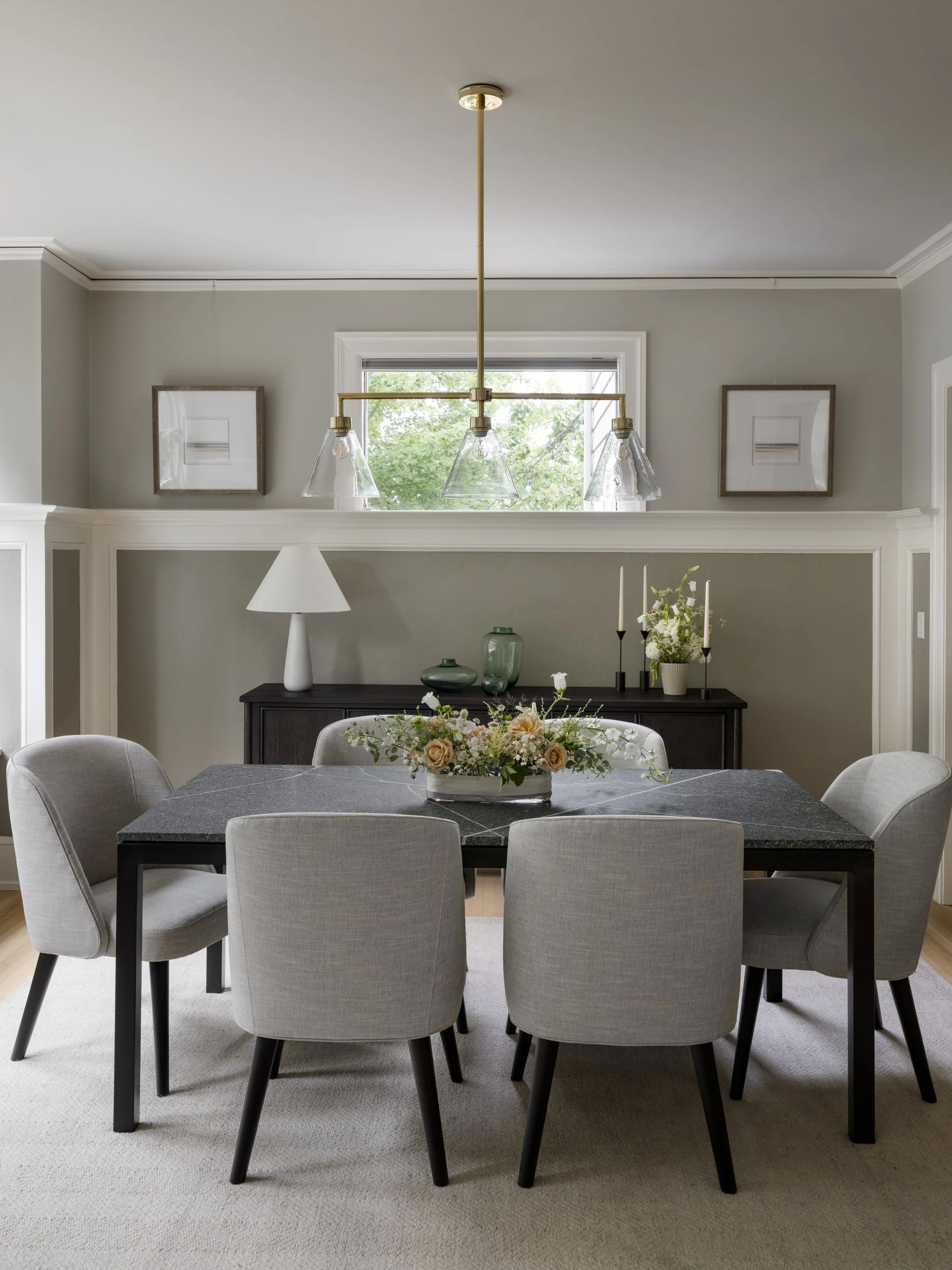 A dining room with a black table, six light gray upholstered chairs, a floral centerpiece, a white table lamp, and decorative vases and candles on a black sideboard. There are framed pictures on the wall and a window showing greenery outside.