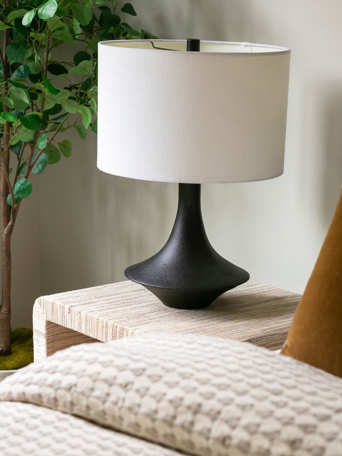 A black ceramic table lamp with a white fabric lampshade sitting on a beige textured side table in a living room, with a green plant in the background.