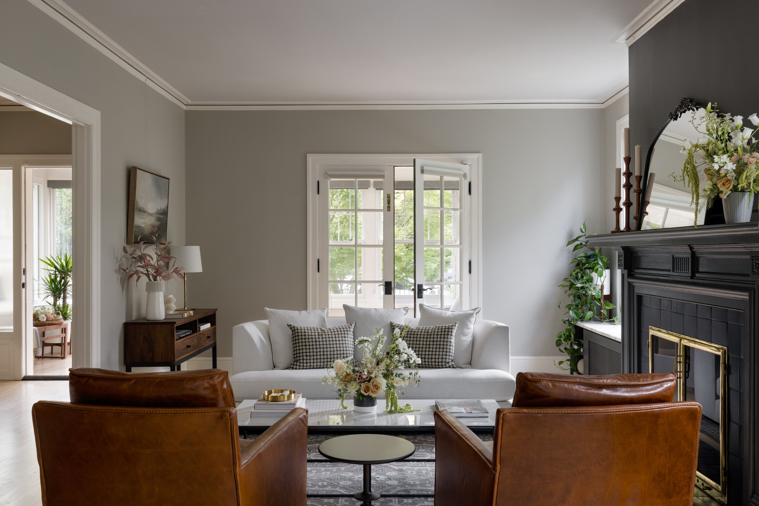 A cozy living room with a white sofa, brown leather armchairs, a black mantel fireplace decorated with a mirror, flowers, and candles, a wooden media console, large windows with greenery outside, and a vase of flowers on the coffee table.