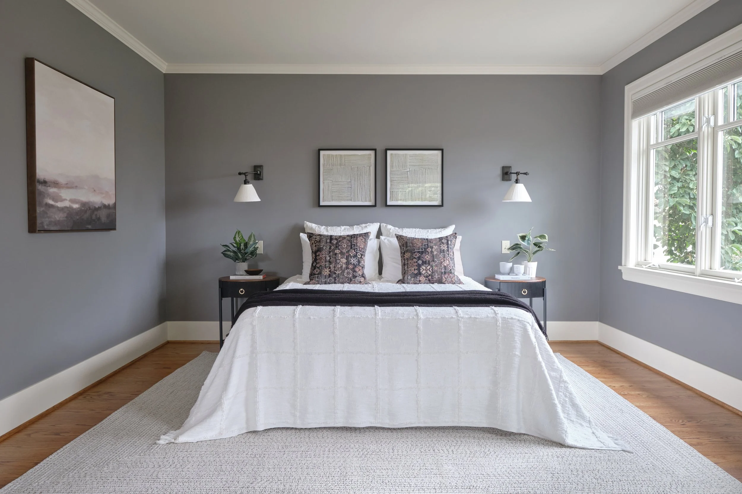 A neatly made bed with white bedding and decorative pillows in a bedroom with gray walls, artwork above the headboard, nightstands with plants, and natural light coming through a window.