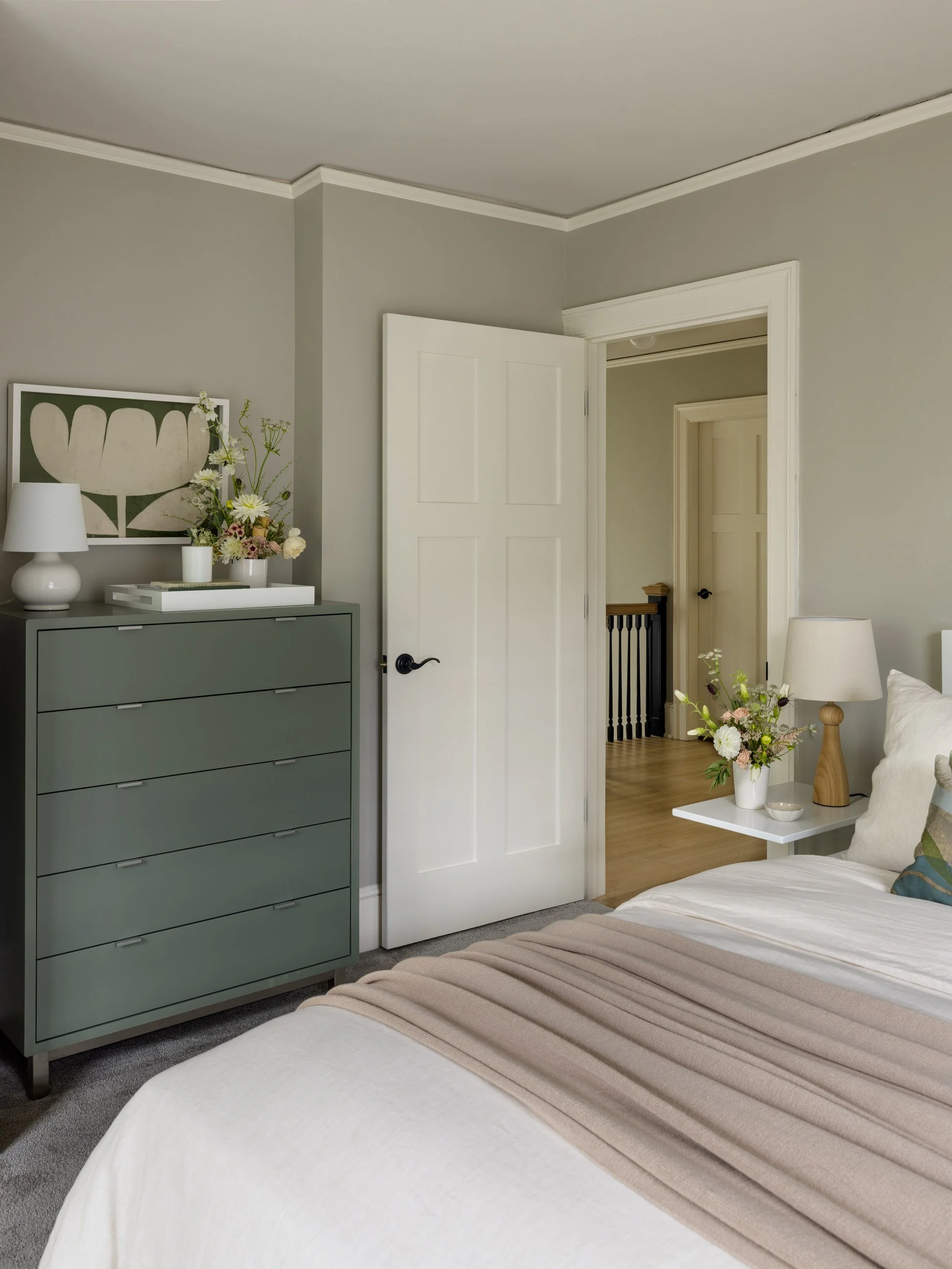 A bedroom with a gray dresser topped with white and pink flowers, a white table lamp, and a small white tray, next to a bed with beige and blue pillows and a beige blanket, with a white door, beige walls, and hardwood floors.