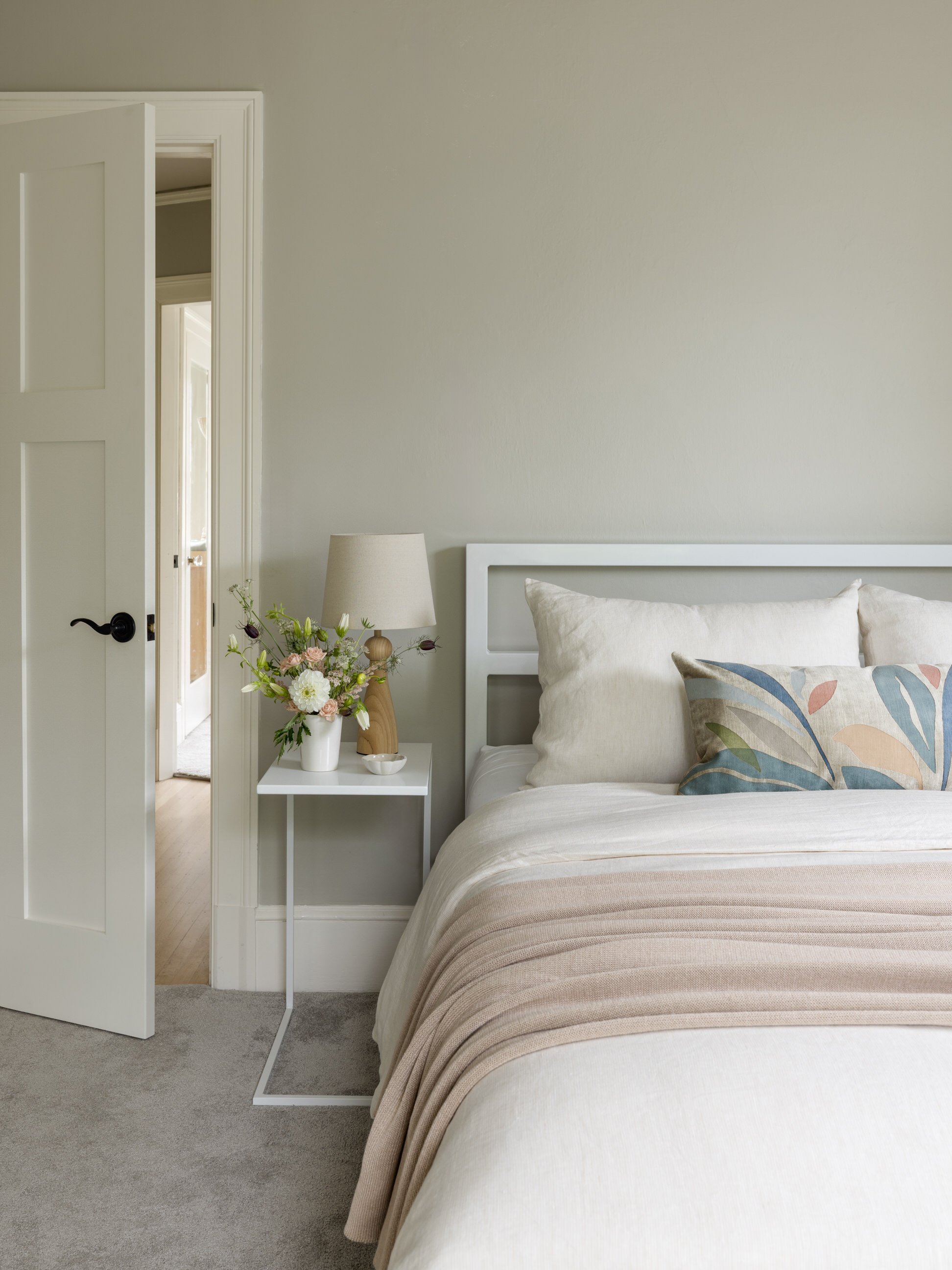 A bedroom with a bed featuring beige bedding and a decorative pillow with a leaf pattern. There is a white nightstand with a table lamp and a vase of flowers beside the bed. An open door reveals another room.