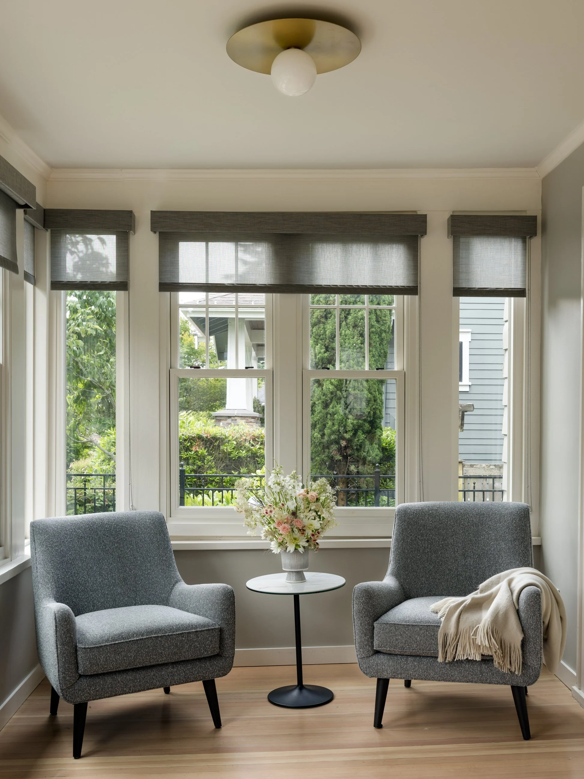 A cozy sitting area with two grey armchairs around a small round black table. A vase with white and pink flowers is on the table. Large windows with grey window treatments and scenic greenery outside allow natural light to fill the room.