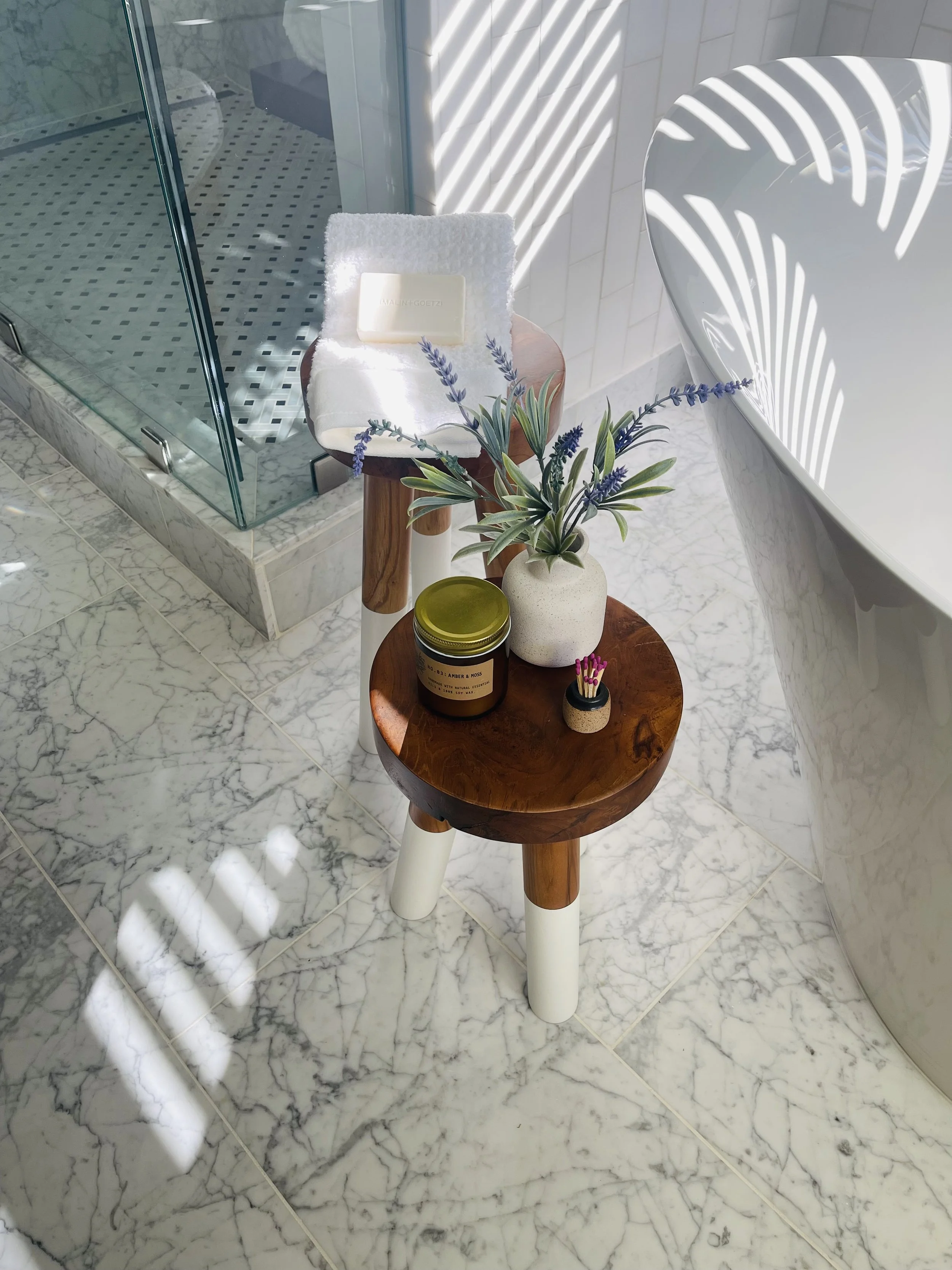 A small round wooden table with white and wooden legs, holding a potted plant with purple flowers, a brown jar labeled 'Amber & Moss,' a small container with matches, and a white towel with a soap bar on top. The table is next to a white bathtub, wit