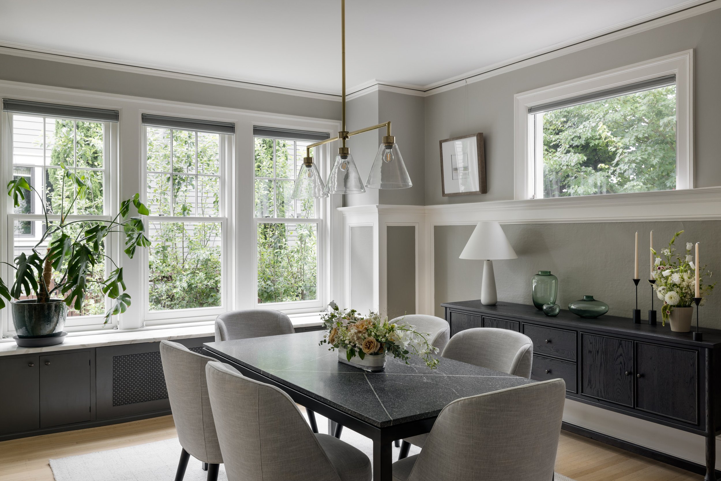 A dining room with large windows letting in natural light, decorated with vases of flowers and plants, and a modern light fixture hanging above the dining table.