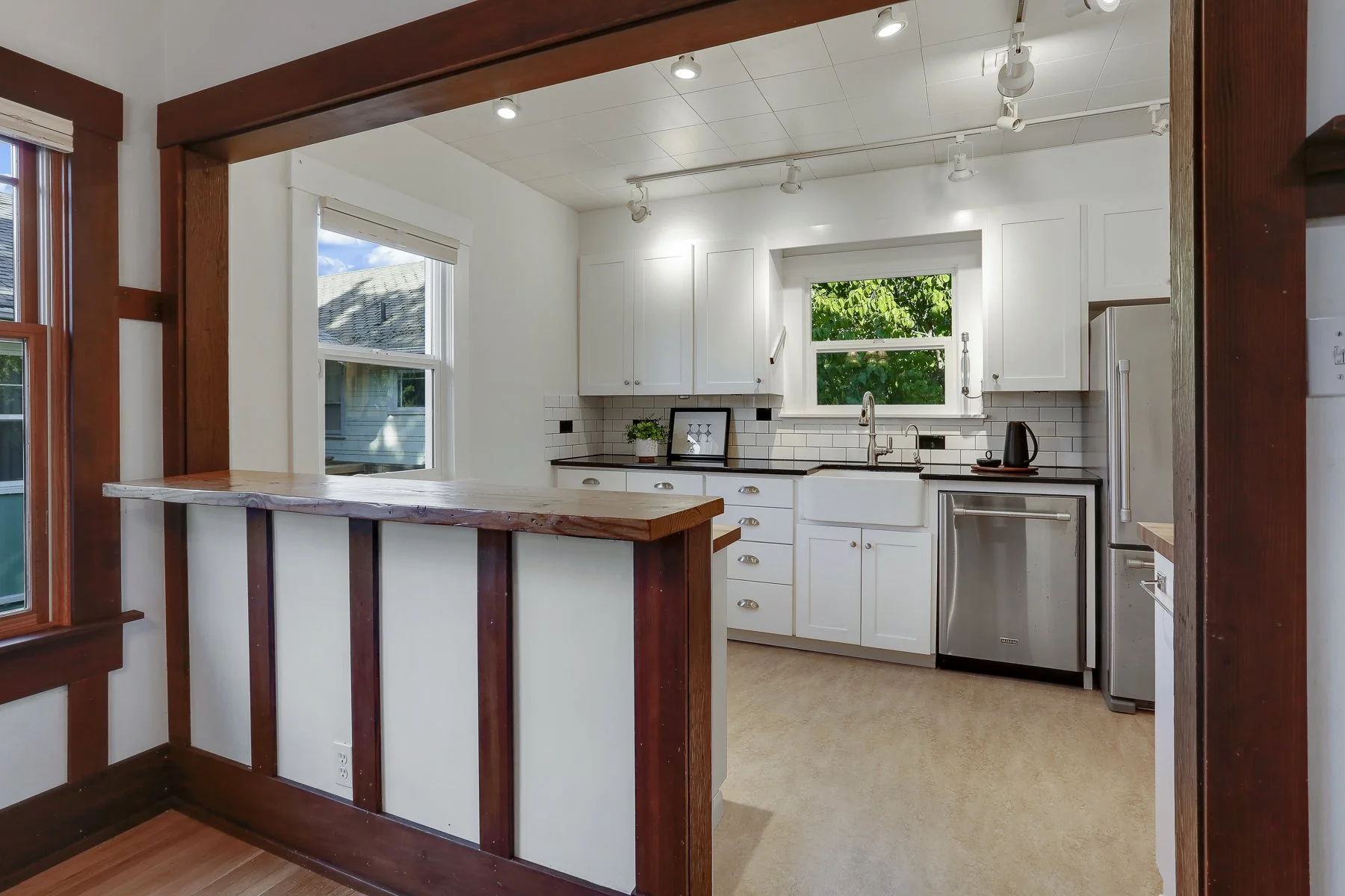 Kitchen with white cabinets, black countertop, stainless steel dishwasher, window showing green trees, and a small bar area with a wooden countertop.