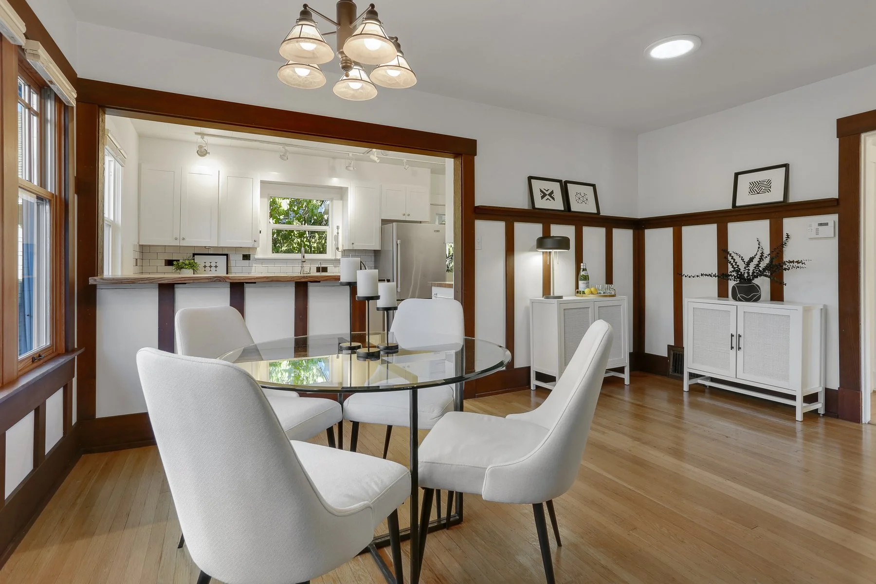 A dining room with a glass-top table and four white upholstered chairs. In the background, a kitchen with white cabinets, a window, and a stainless steel refrigerator. Decor includes black-and-white framed artwork, a black table lamp, and decorative 