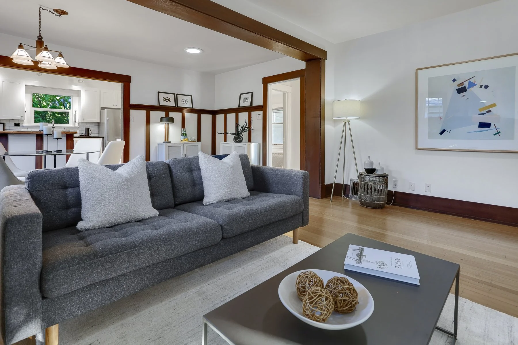 Living room with gray sofa, white pillows, modern art on the wall, wooden floors, and a coffee table with decorative balls.