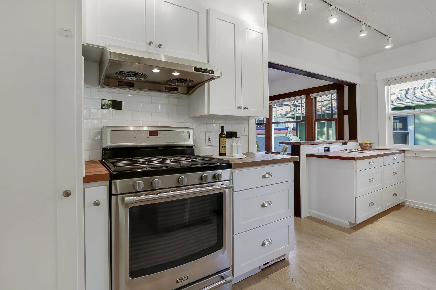 A modern kitchen with white cabinets, a stainless steel oven and stove, a white tiled backsplash, and large windows letting in natural light, with a view of a backyard.