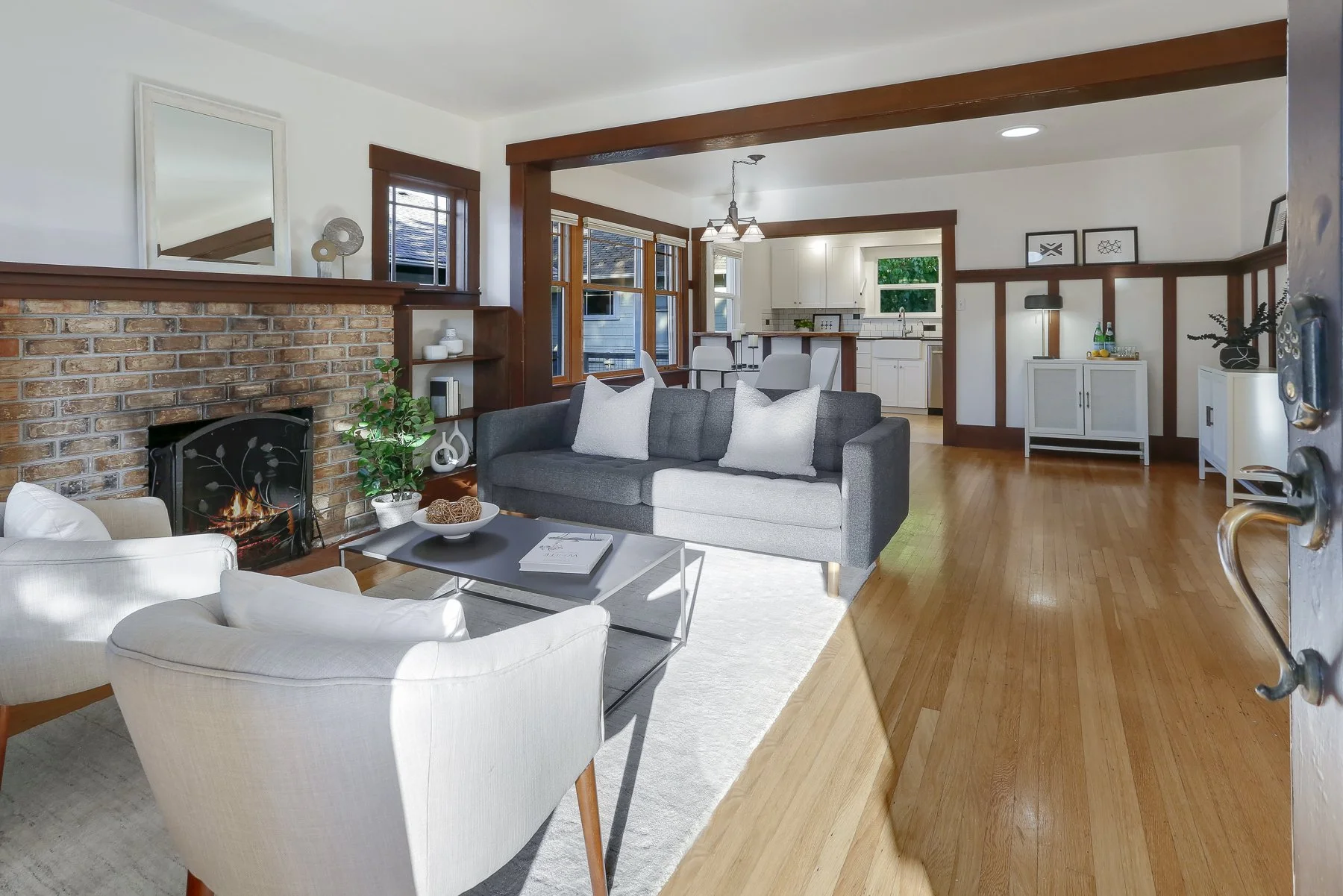 Living room with a brick fireplace, grey sofa with white pillows, white armchair, coffee table with decorative items, and a view into the kitchen with white cabinets and wooden accents.