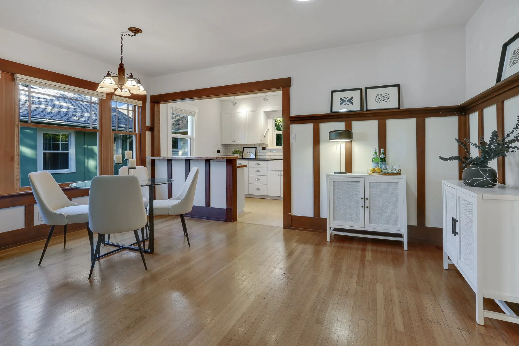 Living room with hardwood floors, white walls with wood trim, a round dining table with four white chairs, large windows, and a view of the outside. In the background, a kitchen with white cabinetry and a small bar area. There is a white cabinet with