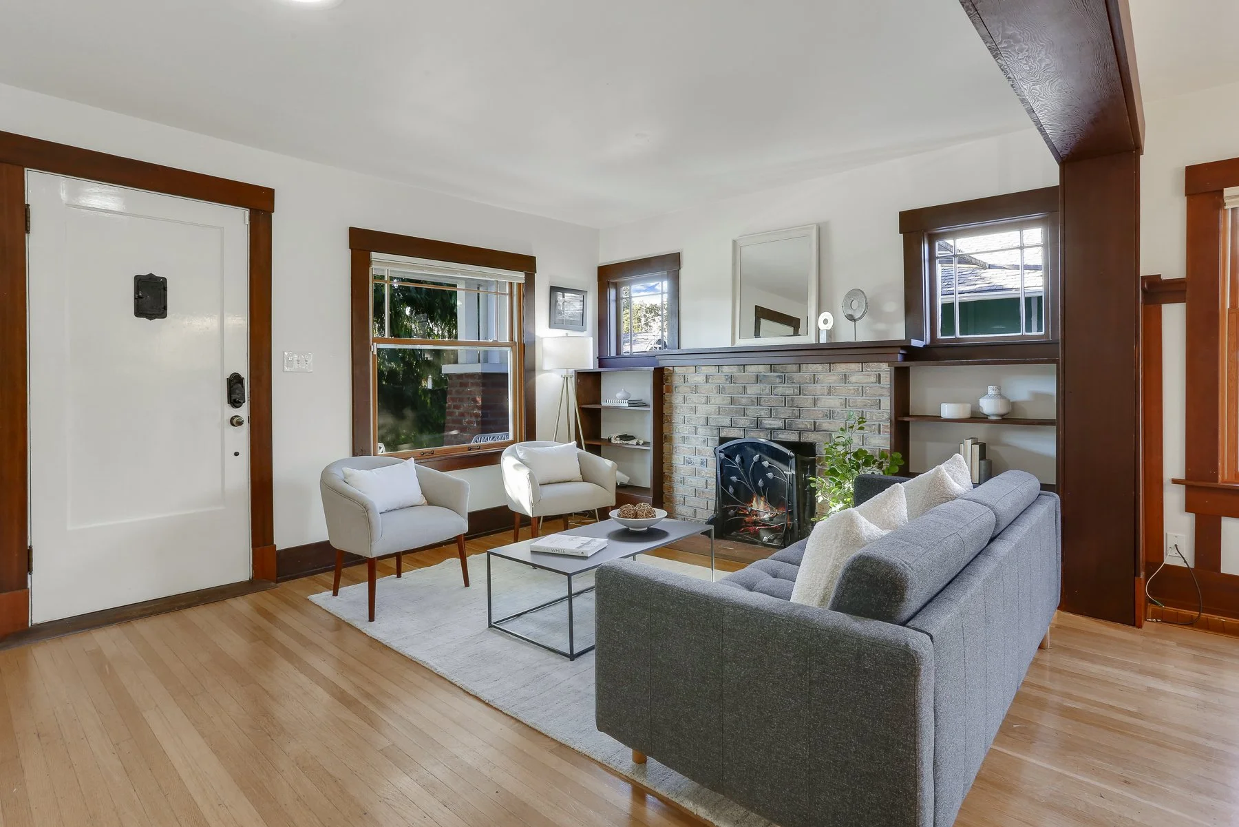 Living room with white walls, wooden trim, a brick fireplace, a gray sofa, white armchairs, a coffee table, and shelves with decorative items.