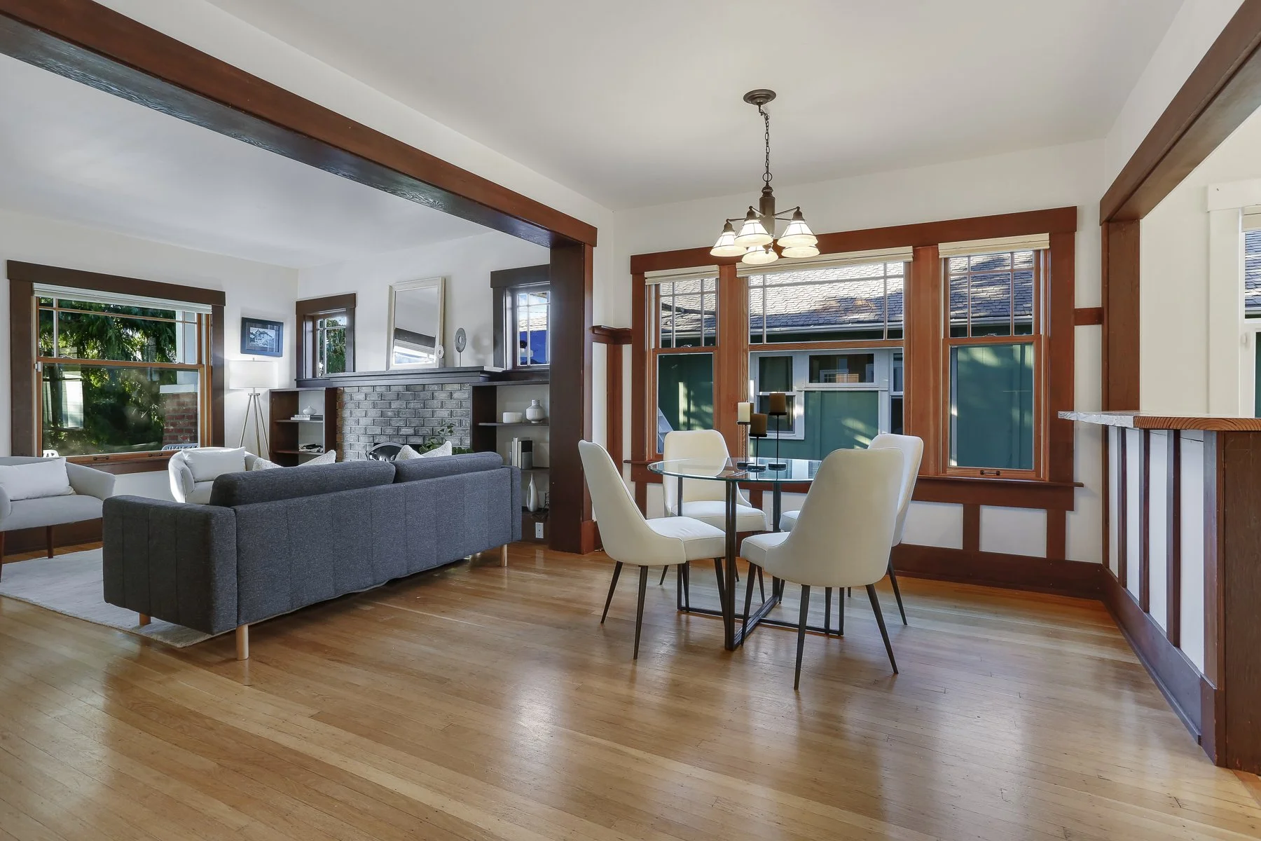 Open living and dining room with wooden trim, large windows, hardwood floors, gray sofa, white armchairs, brick fireplace, and black dining table with white chairs, lit by a chandelier.