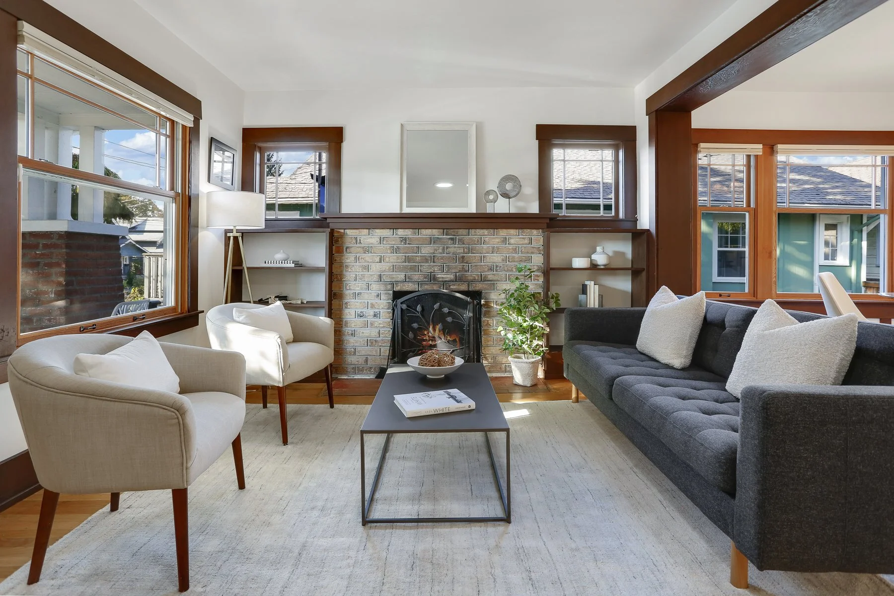 A living room with large windows, a brick fireplace, and a mix of modern and vintage furniture, including white and black sofas, a coffee table, and decorative items.
