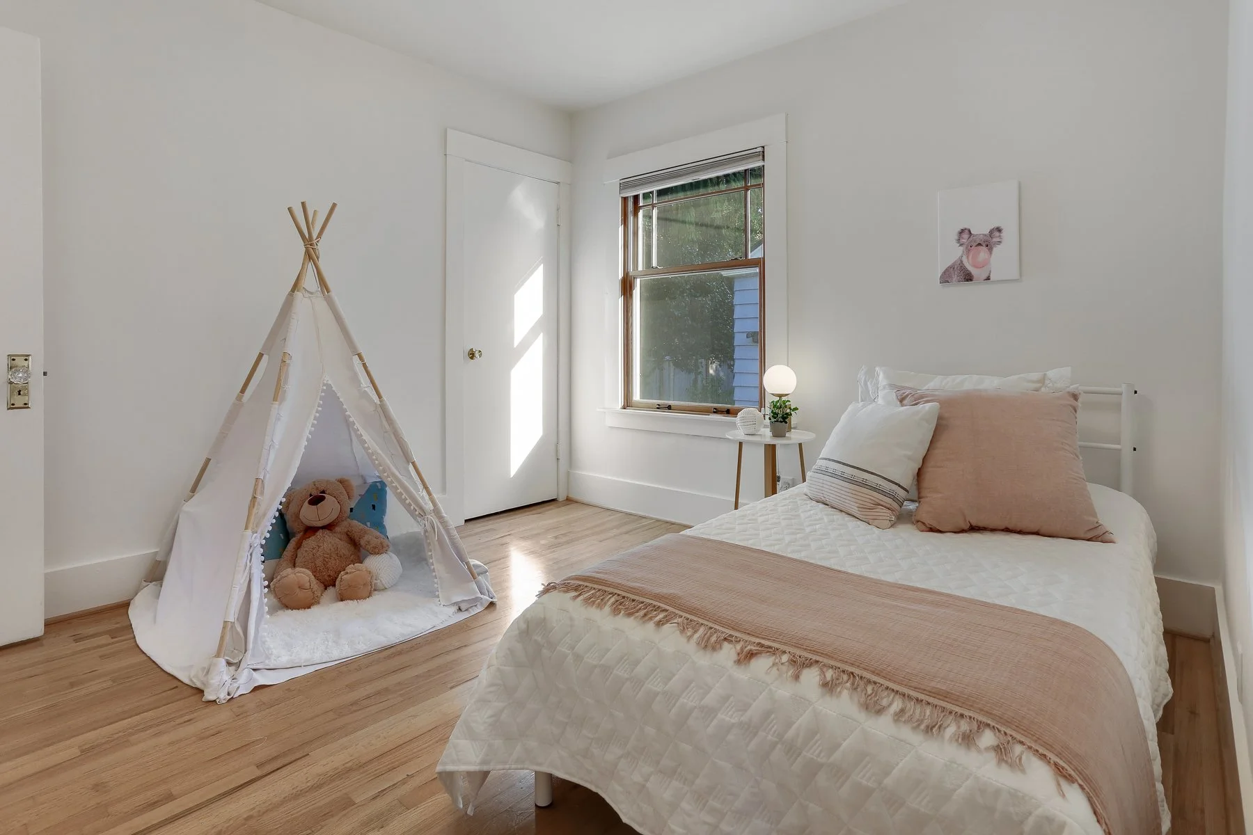 A cozy bedroom with light-colored walls, hardwood flooring, a bed with a peach and white quilt, and a teddy bear inside a white fabric teepee with wooden poles. Next to the bed is a small white side table with a lamp, a plant, and decorative items. T