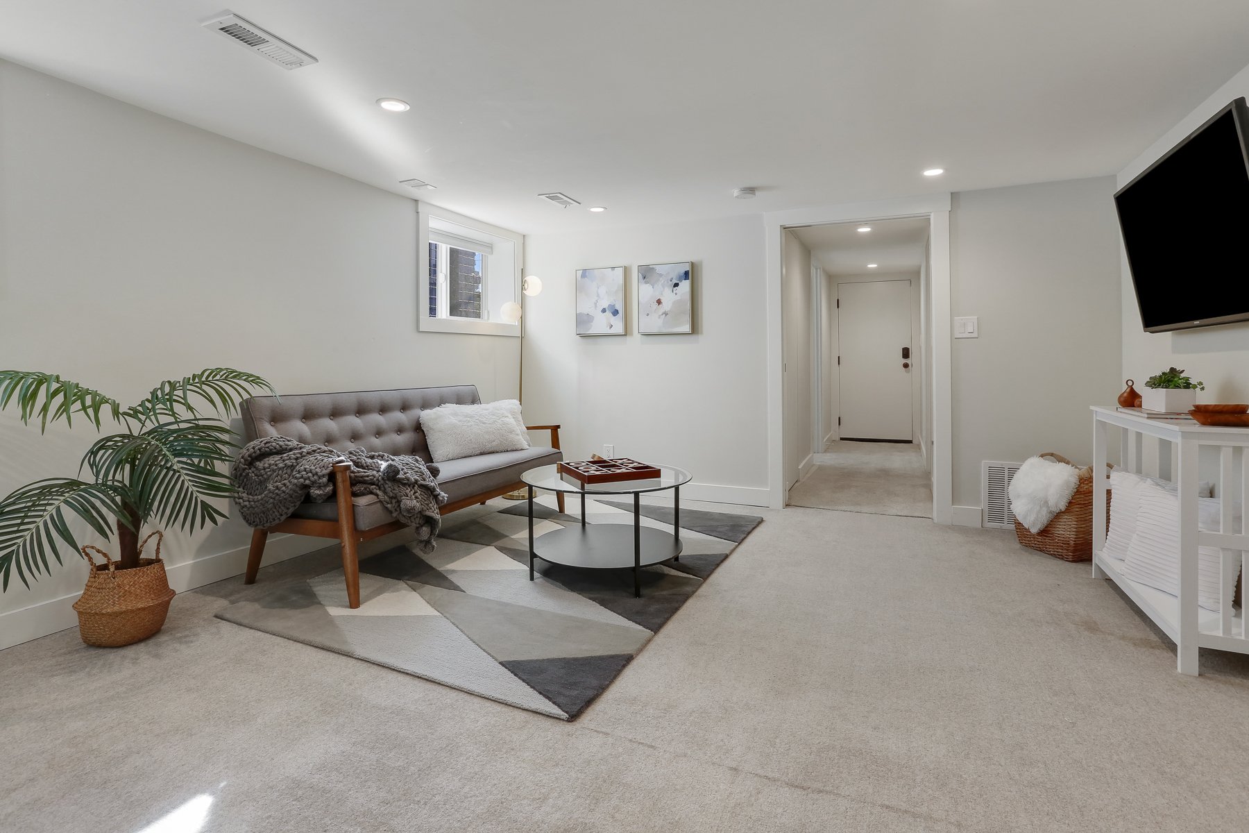 Modern living room with a gray sofa, a round glass coffee table, a potted plant, wall art, a white console table, wall-mounted TV, and decorative items in a neutral color palette.
